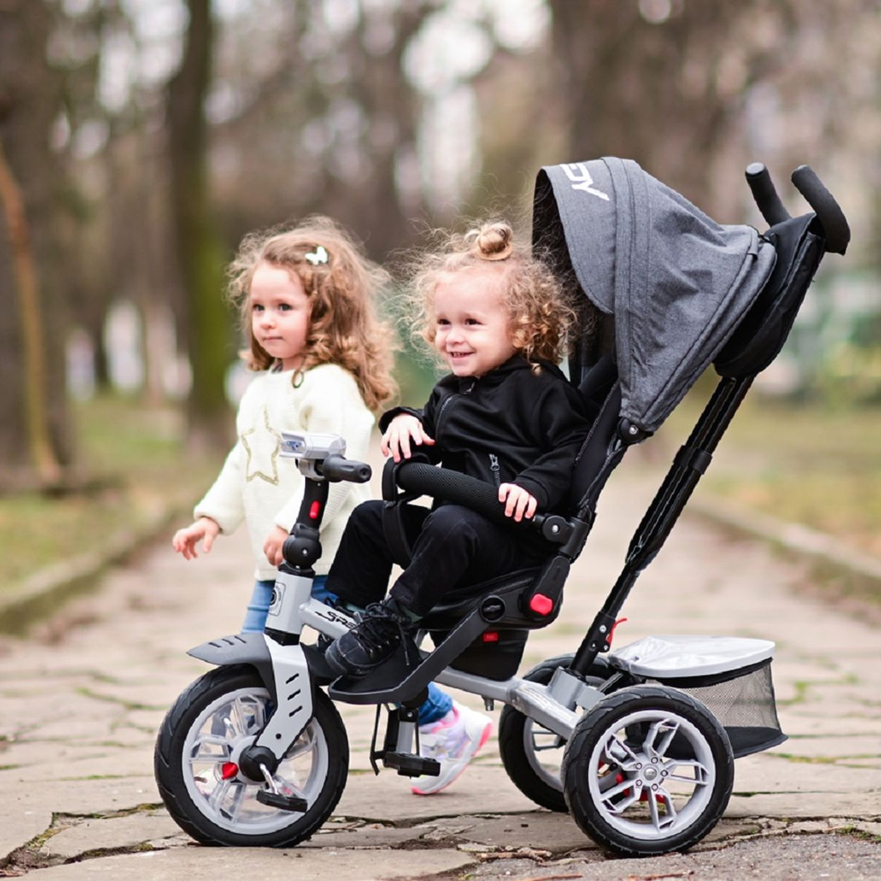 Zwei kleine Mädchen gehen auf einem Weg. Eines fährt in einem grauen Kinderwagen, das andere geht daneben.