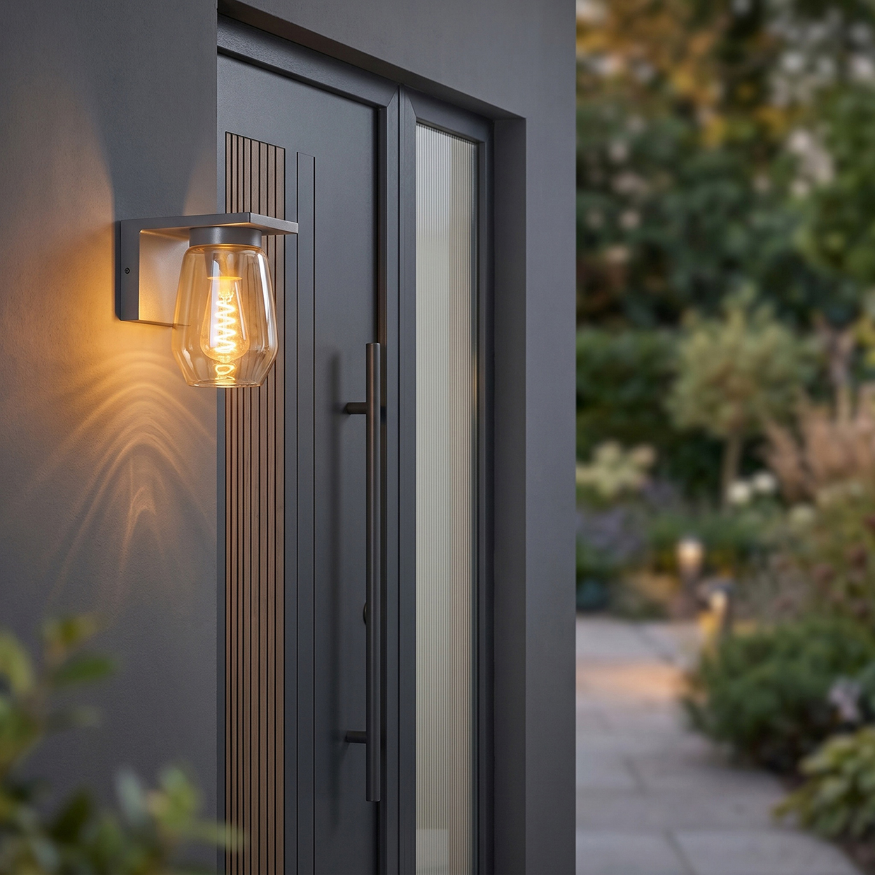 Eine Wandleuchte beleuchtet eine dunkle Haustür mit Glasdetails und einem Garten im Hintergrund.
