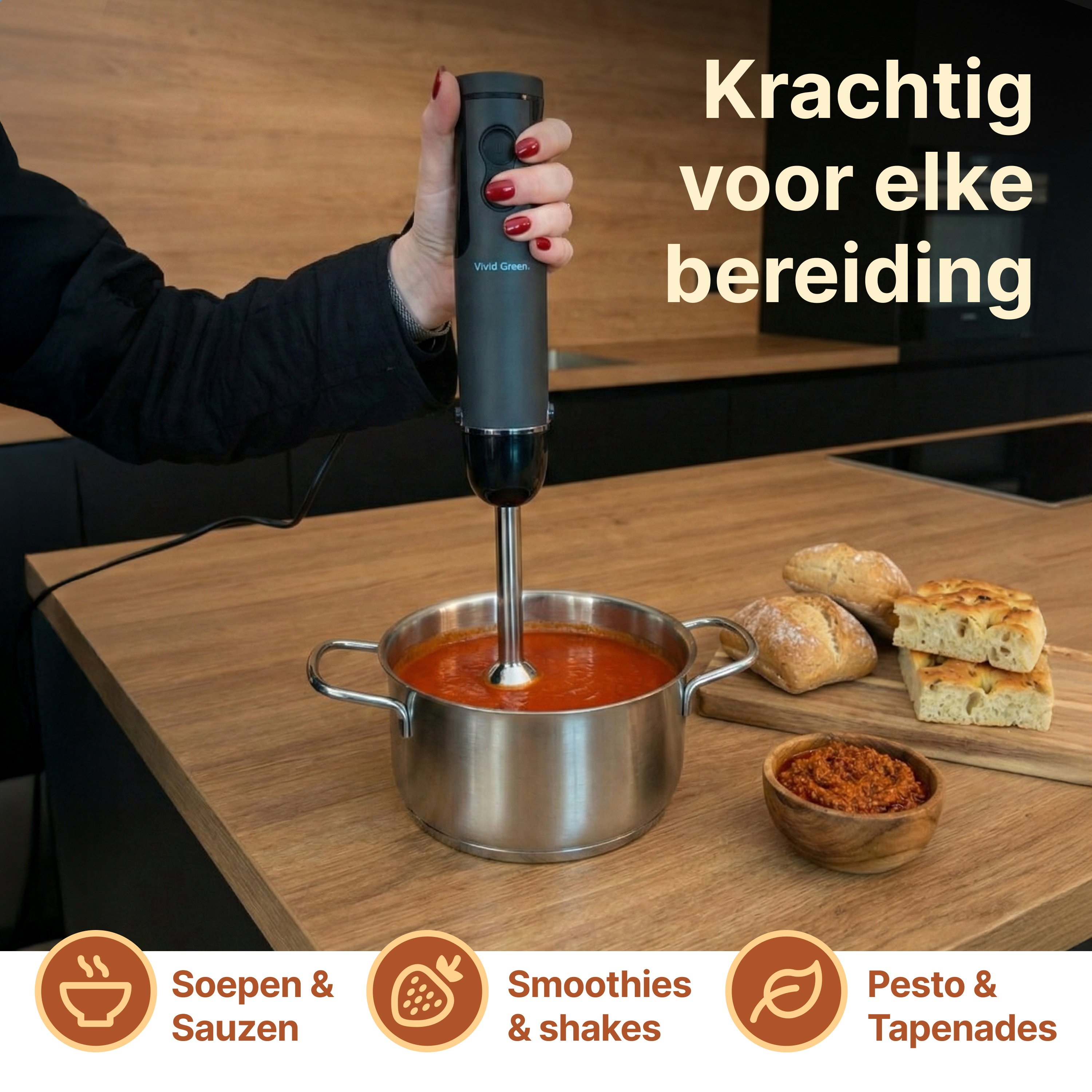 A hand holds an immersion blender over a pot of tomato soup. Bread and dip nearby.
