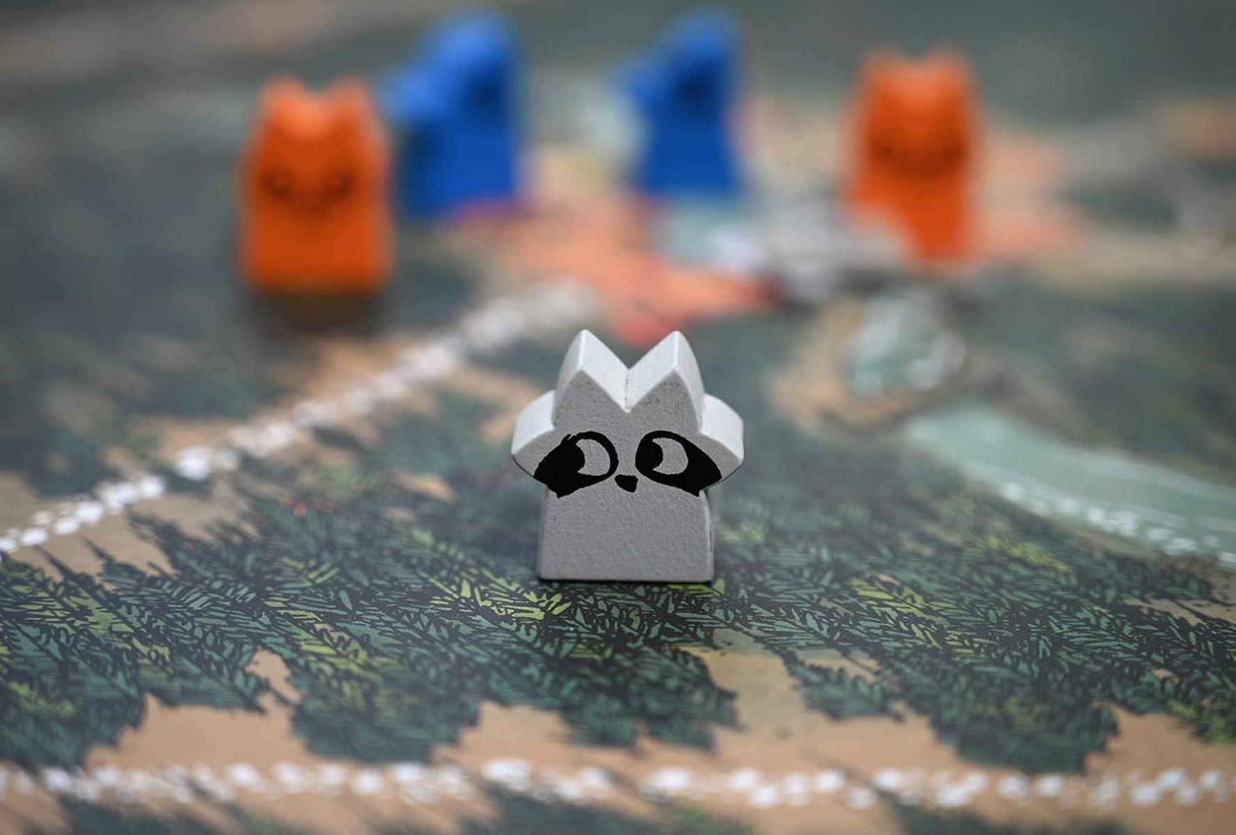Ein graues Waschbär-Spielstein steht im Vordergrund, mit roten und blauen Spielfiguren im Hintergrund.