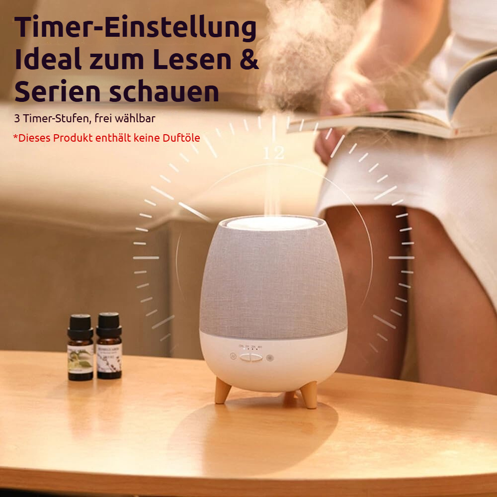 Ein Diffusor mit zwei Ölflaschen auf einem Tisch, im Hintergrund liest eine Person.