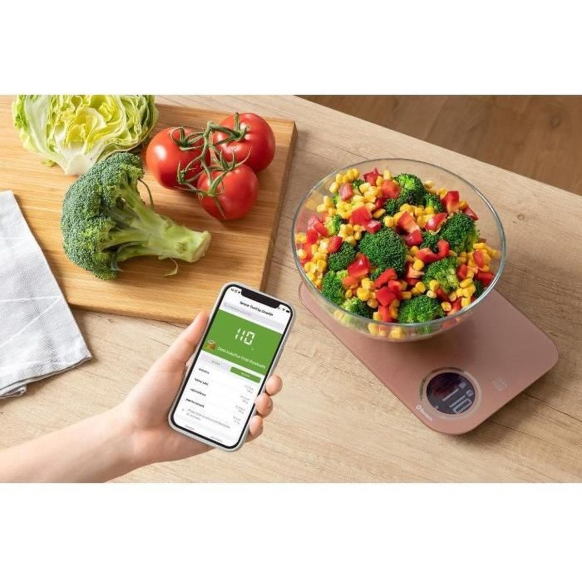Eine Waage mit einem Gemüsesalat, daneben Gemüse und ein Smartphone.