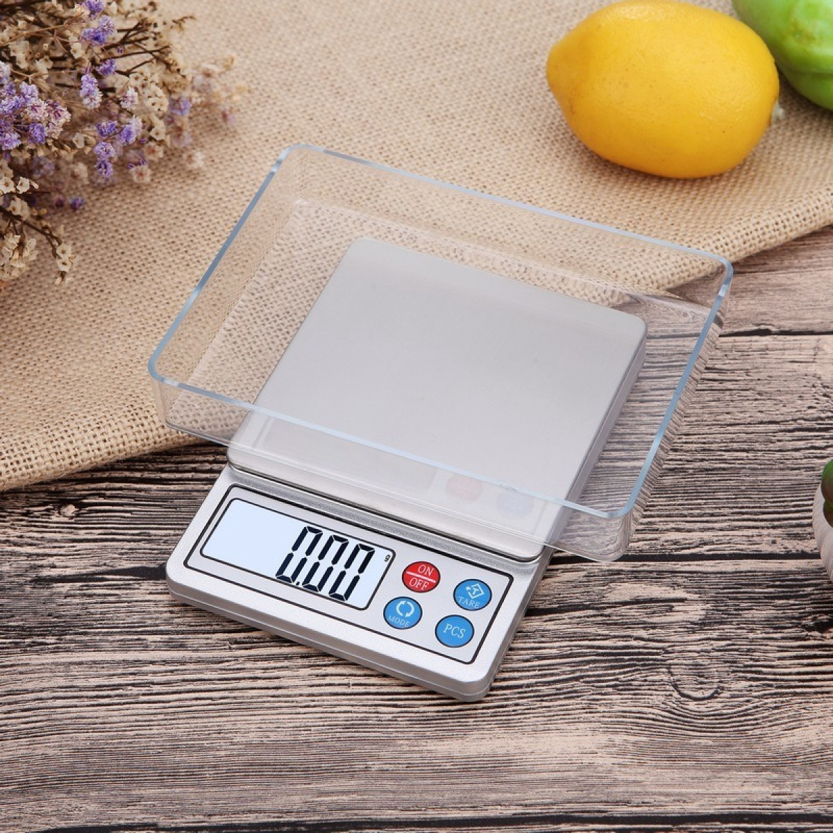 Eine digitale Küchenwaage mit transparenter Schale steht auf Holz mit Trockenblumen und Obst.