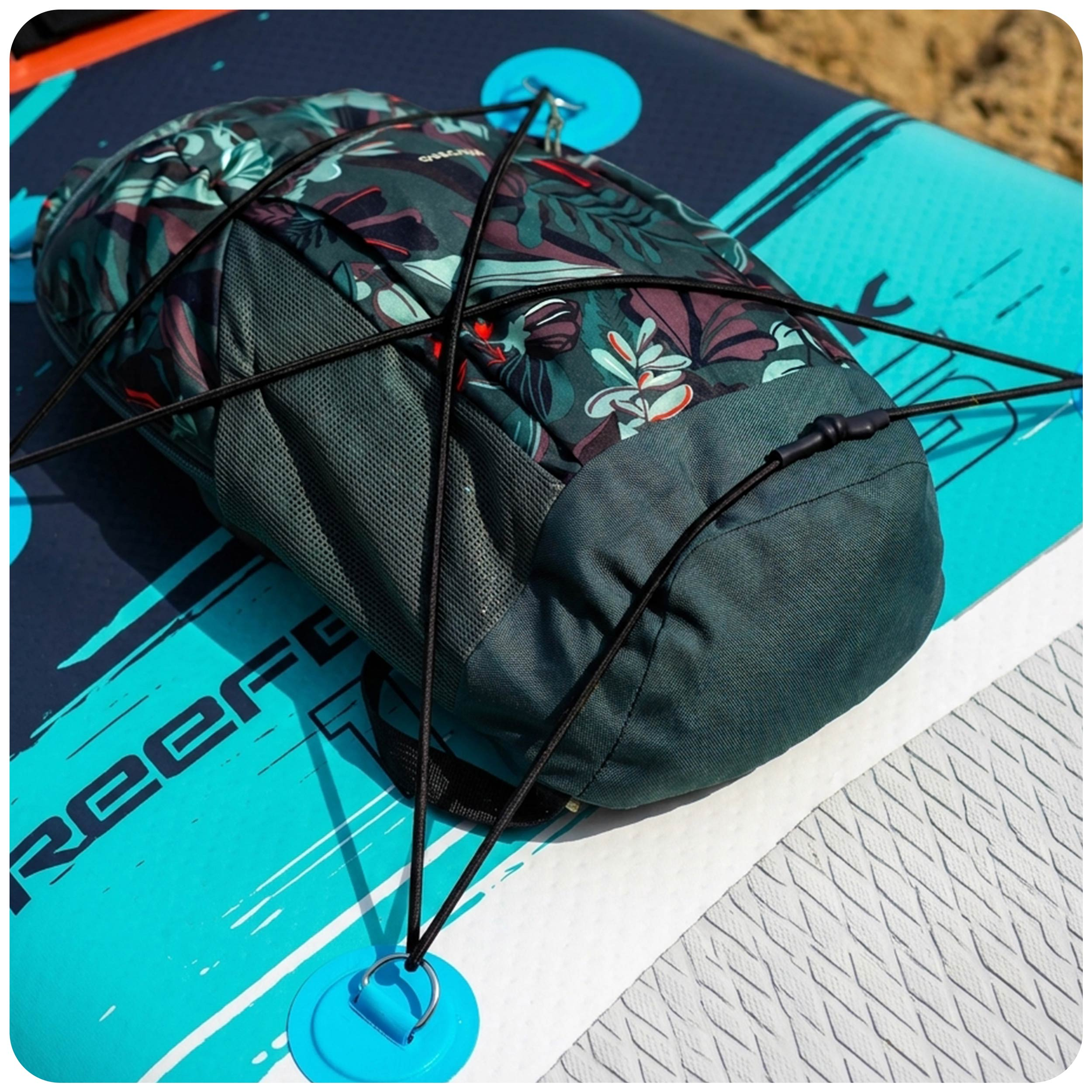 Plecak w kwiatowy wzór zabezpieczony czarnymi linkami bungee na niebieskim paddleboardzie.
