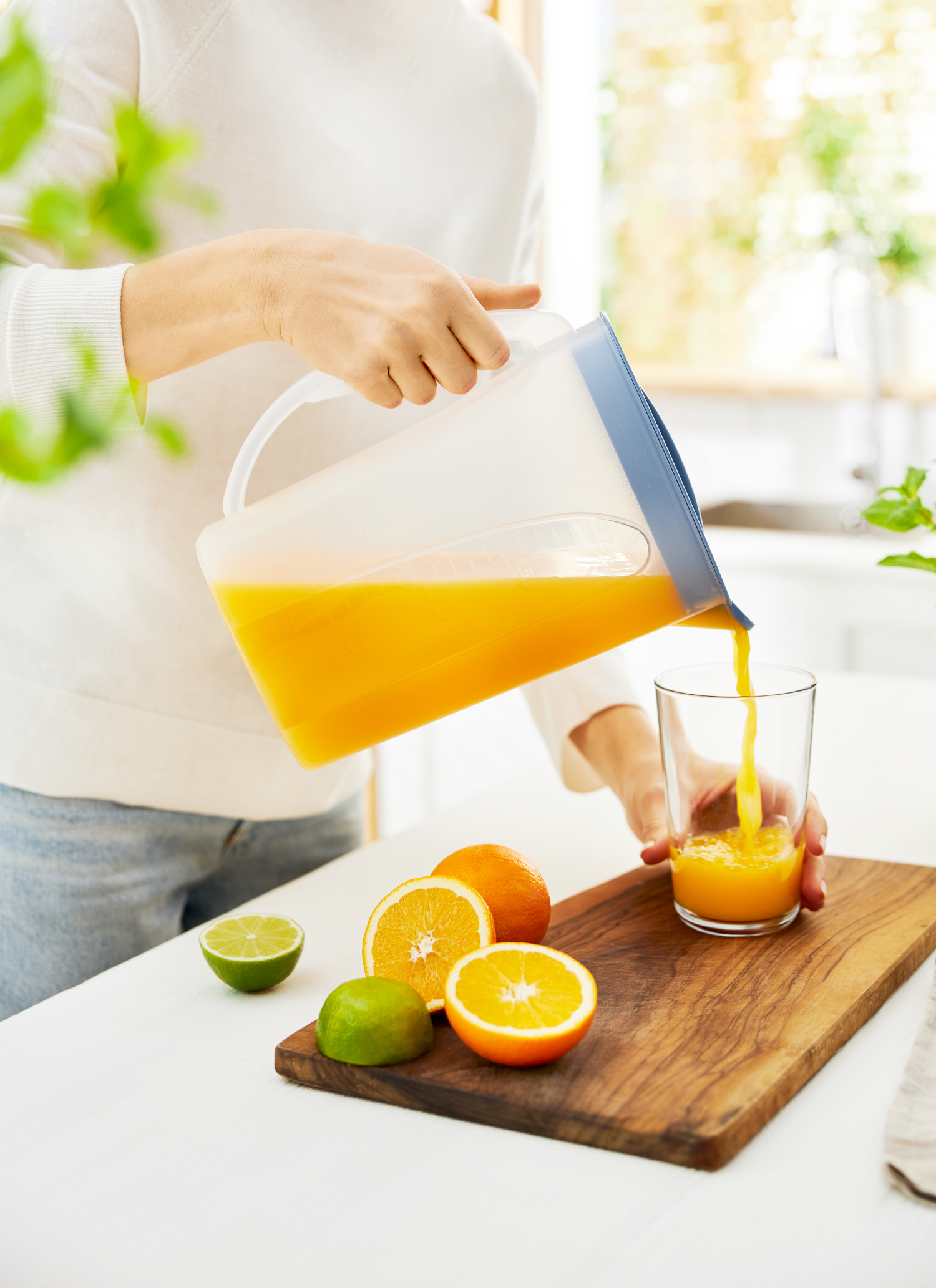 Osoba nalewa sok pomarańczowy z dzbanka do szklanki. Pomarańcze i limonki są w pobliżu.