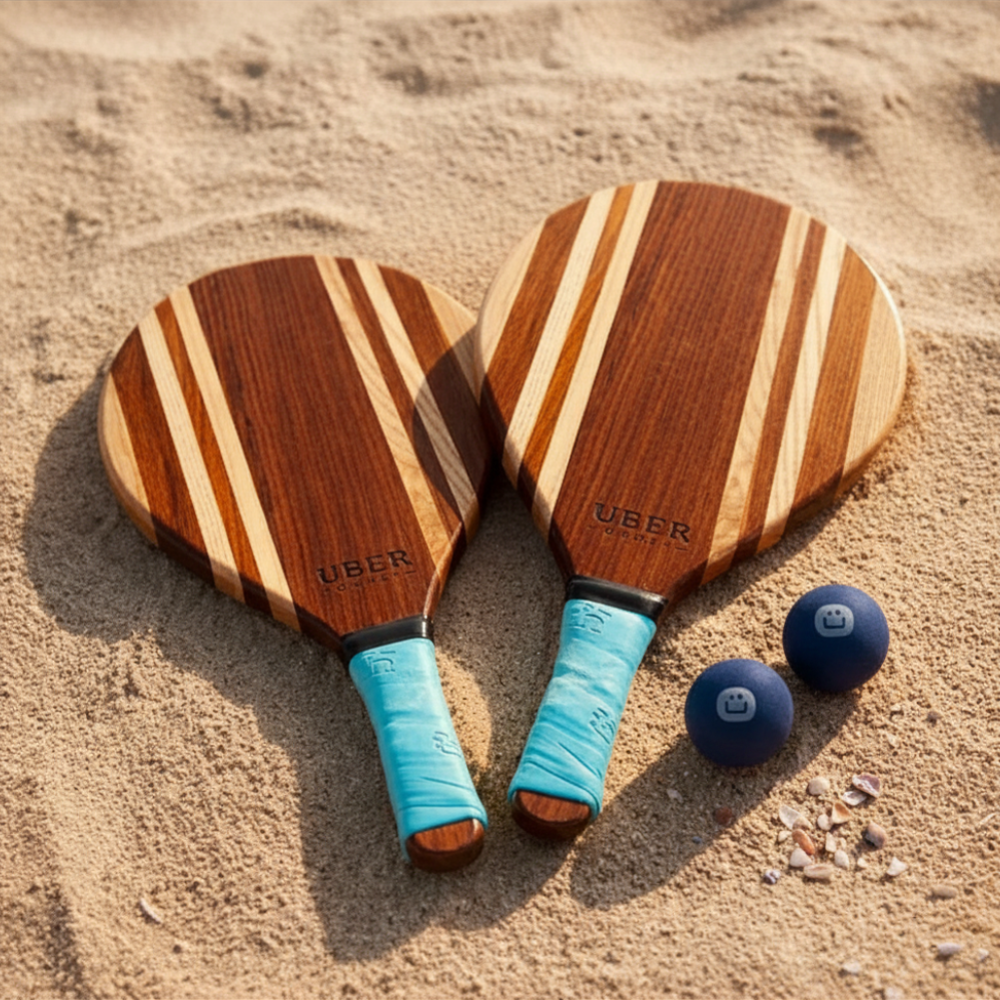 Zwei Holz-Strandschläger mit blauen Griffen und zwei blauen Bällen liegen im Sand.