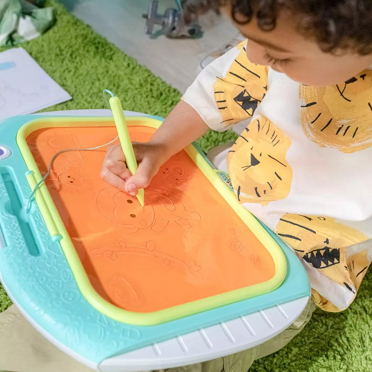 Un bambino disegna su una lavagna magnetica arancione con una penna gialla. Il bambino indossa una maglietta bianca con stampa tigre.