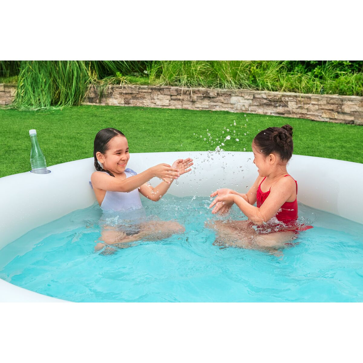 Dos niñas chapotean en una piscina inflable en un jardín. Una lleva un bañador a rayas, la otra uno rojo.