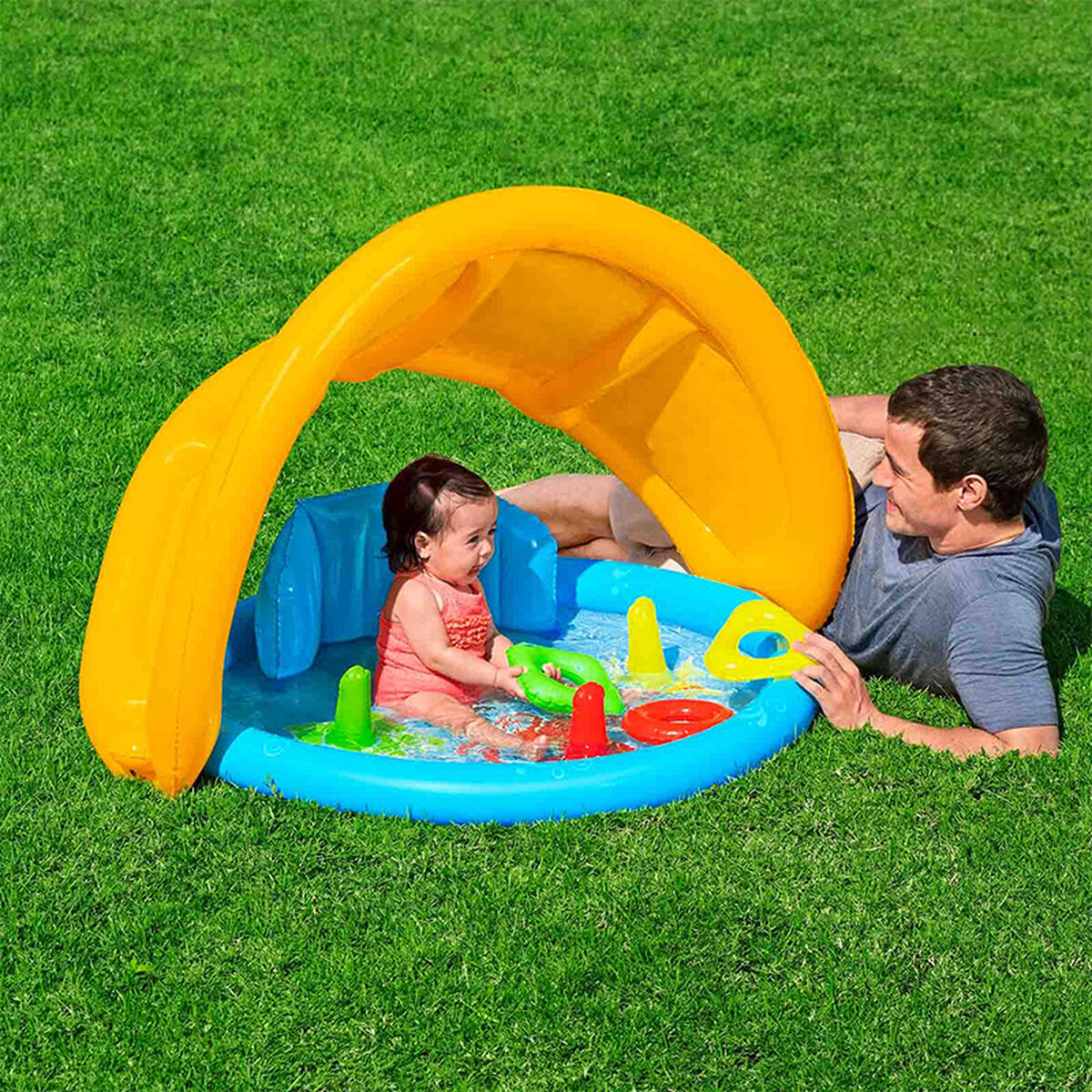 Un hombre yace en el césped observando a un bebé en una piscina infantil bajo un parasol.