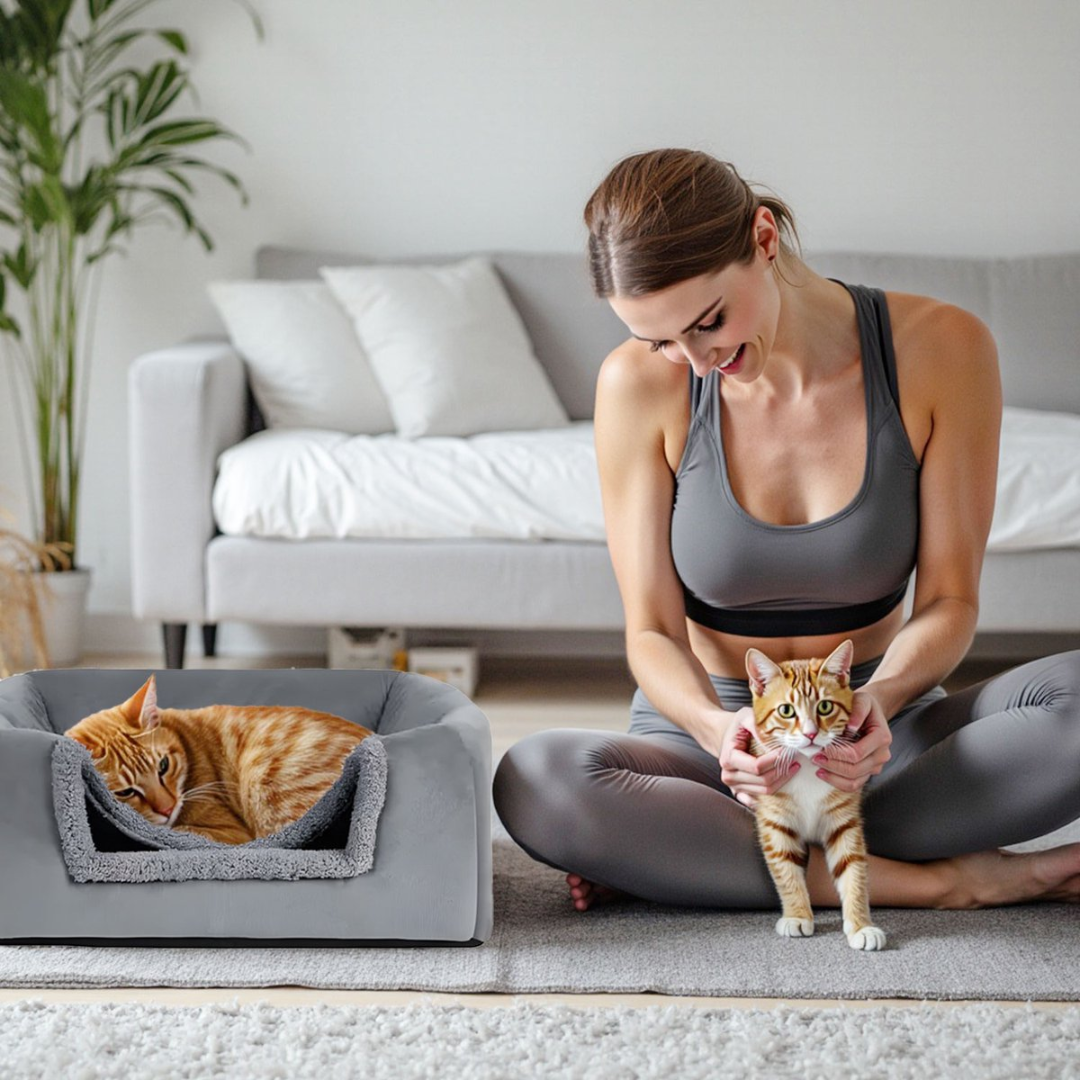 A woman in athletic wear sits on the floor holding a cat. Another cat rests in a pet bed.