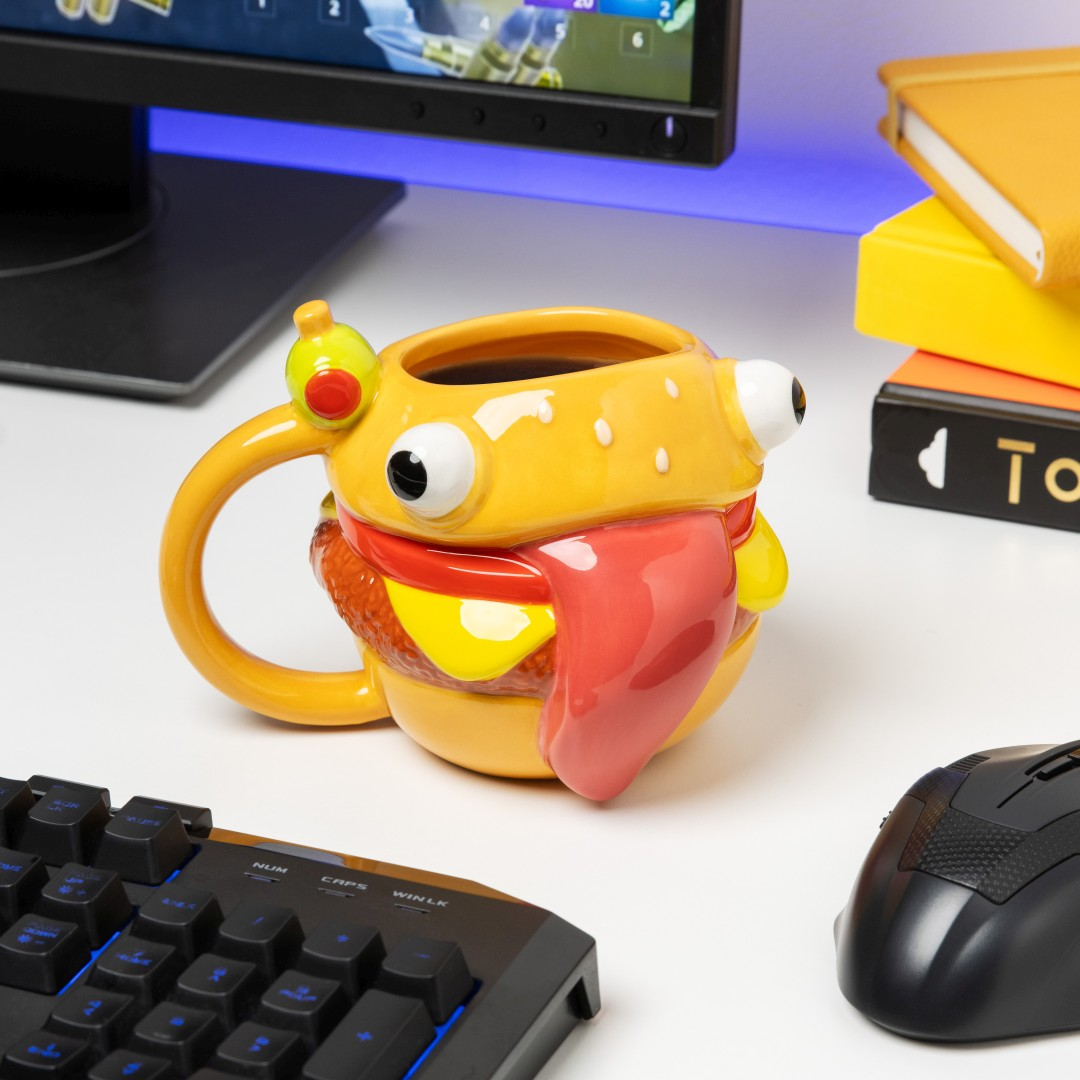 Una taza con forma de hamburguesa, con ojos y lengua, está sobre un escritorio junto a un teclado y ratón.