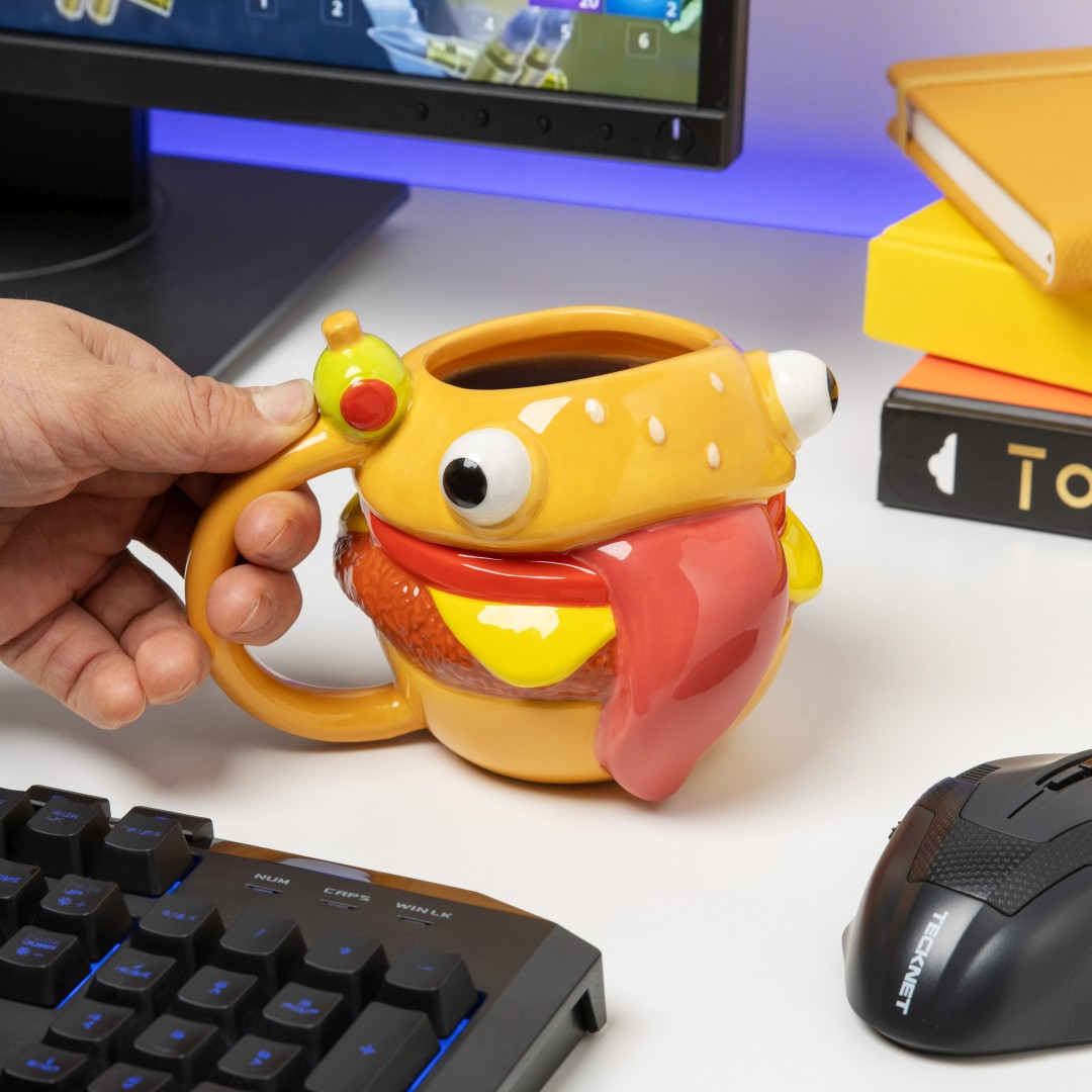 Una mano sostiene una taza con forma de hamburguesa con ojos y lengua colgante junto a un teclado.
