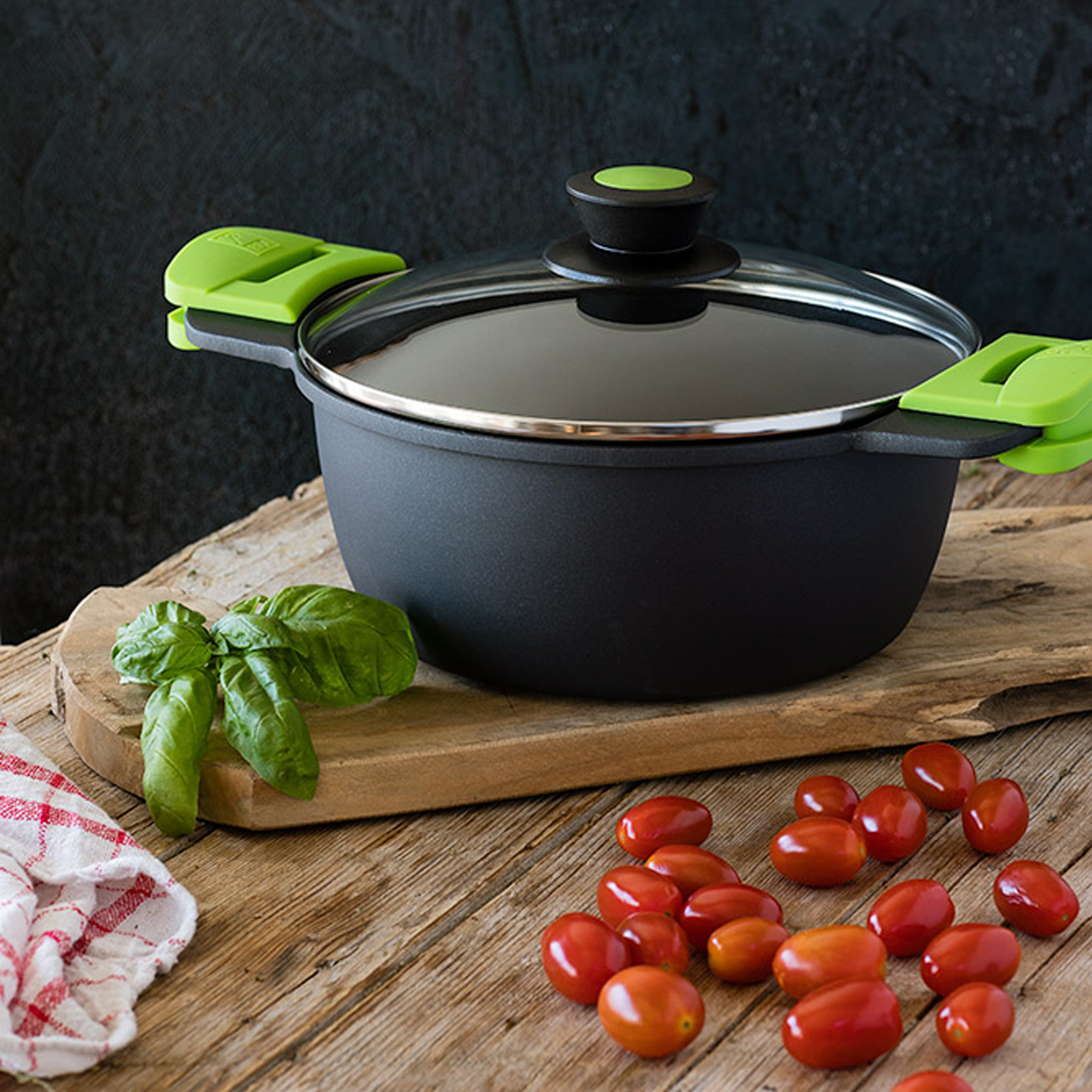 Olla negra con tapa de cristal y asas verdes, junto a albahaca y tomates cherry.