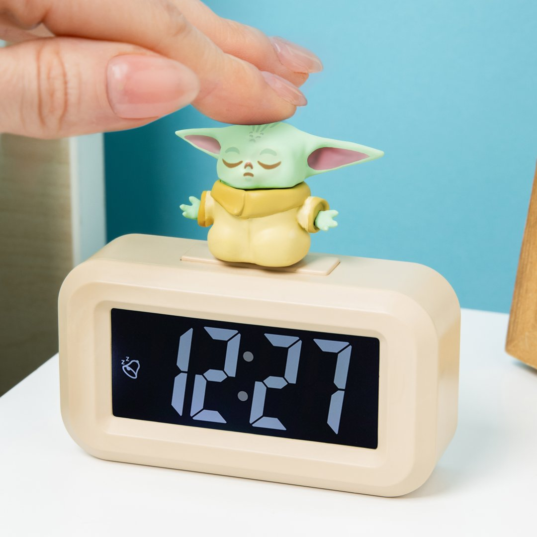 Figurka Baby Yody siedzi na beżowym budziku cyfrowym wyświetlającym 12:27.