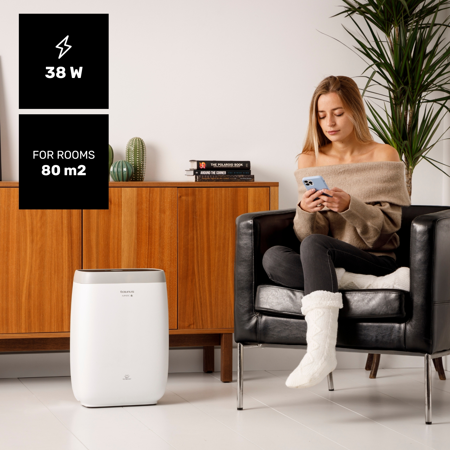Un purificador de aire blanco está al lado de un sillón negro donde se sienta una mujer.
