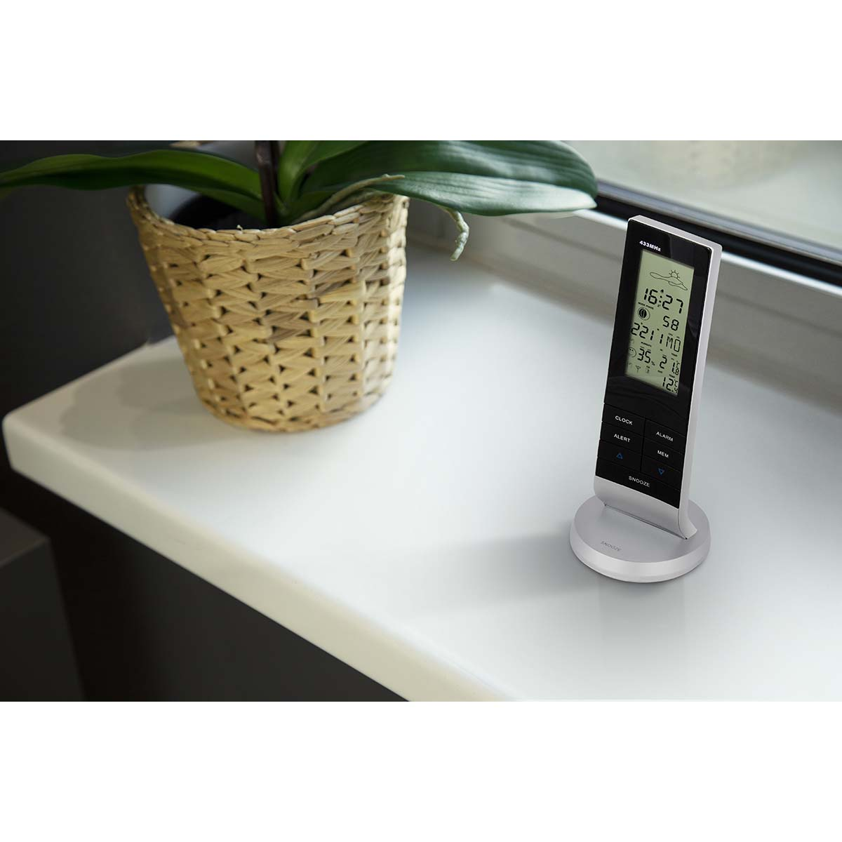 Ein digitales Wetterstation auf einem weißen Sims mit einer Topfpflanze.