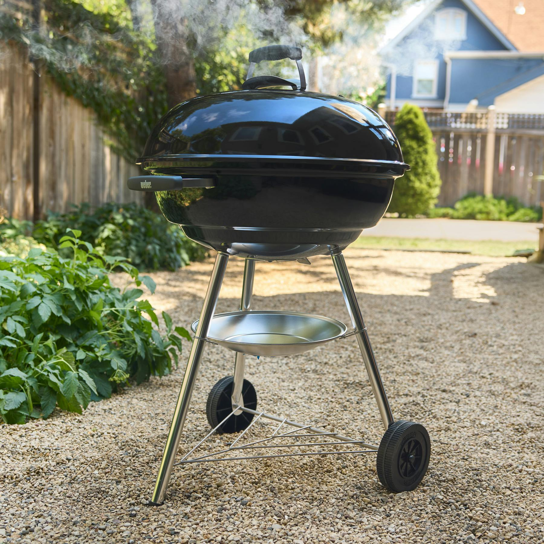 Ein schwarzer Weber Grill mit Deckel, auf dem Kies im Garten steht. Rauch steigt auf.