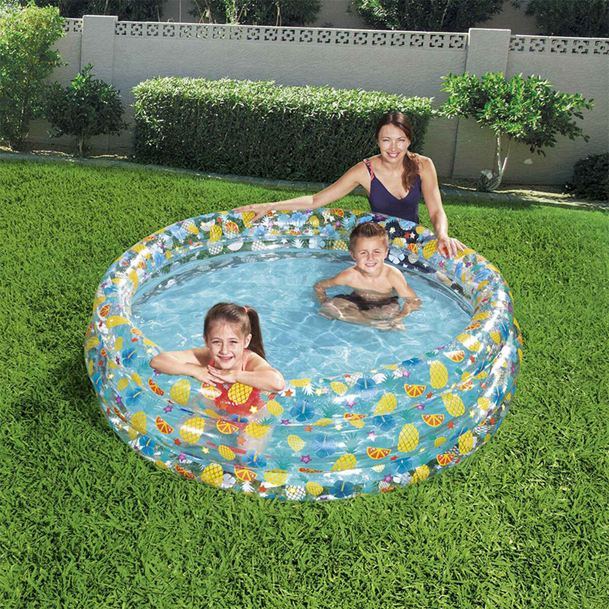 Una mujer y dos niños chapotean en una piscina inflable frutal en un jardín.