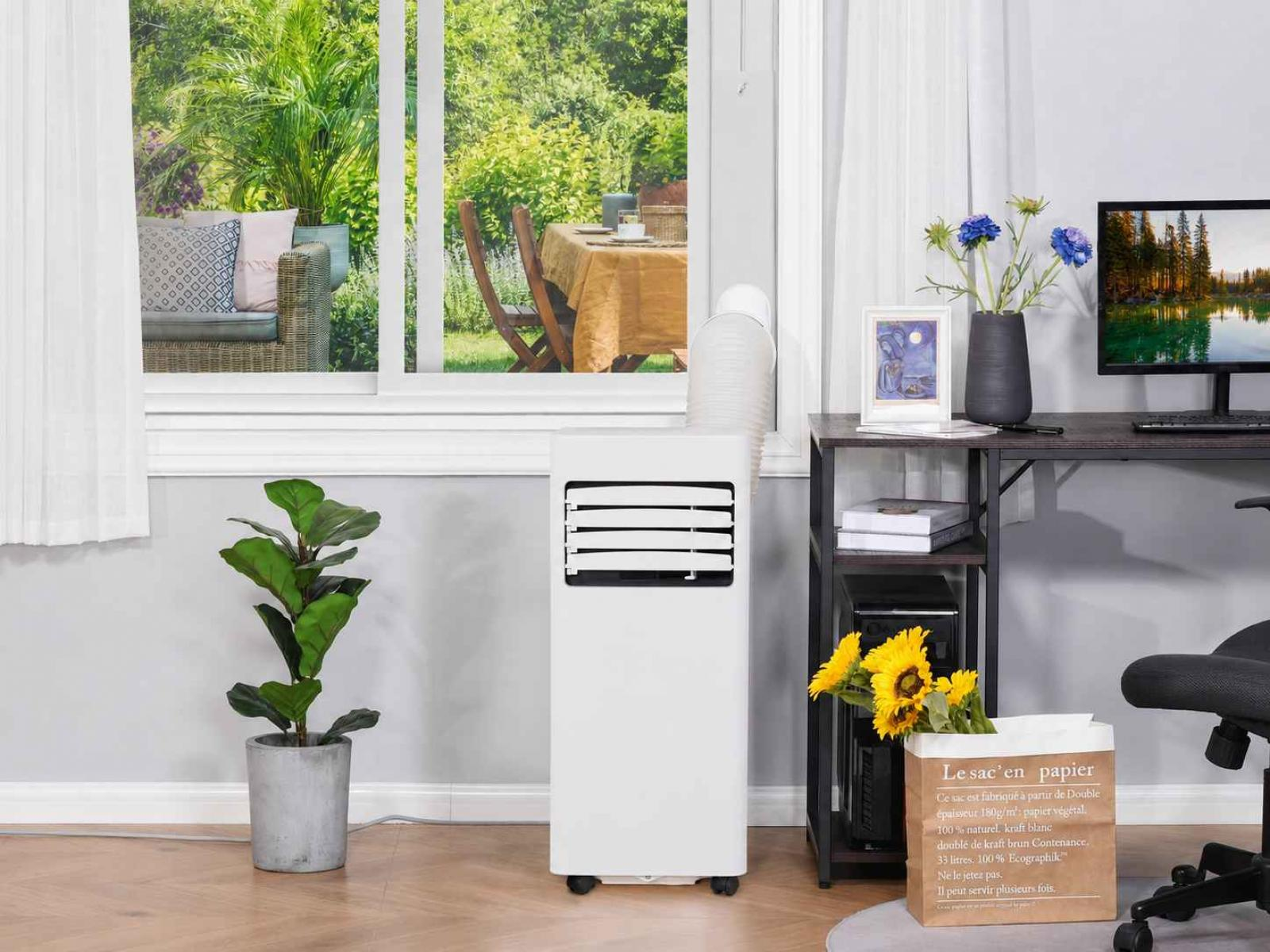 Ein weißer mobiler Klimaanlage steht neben einem Fenster mit Blick auf einen Garten.