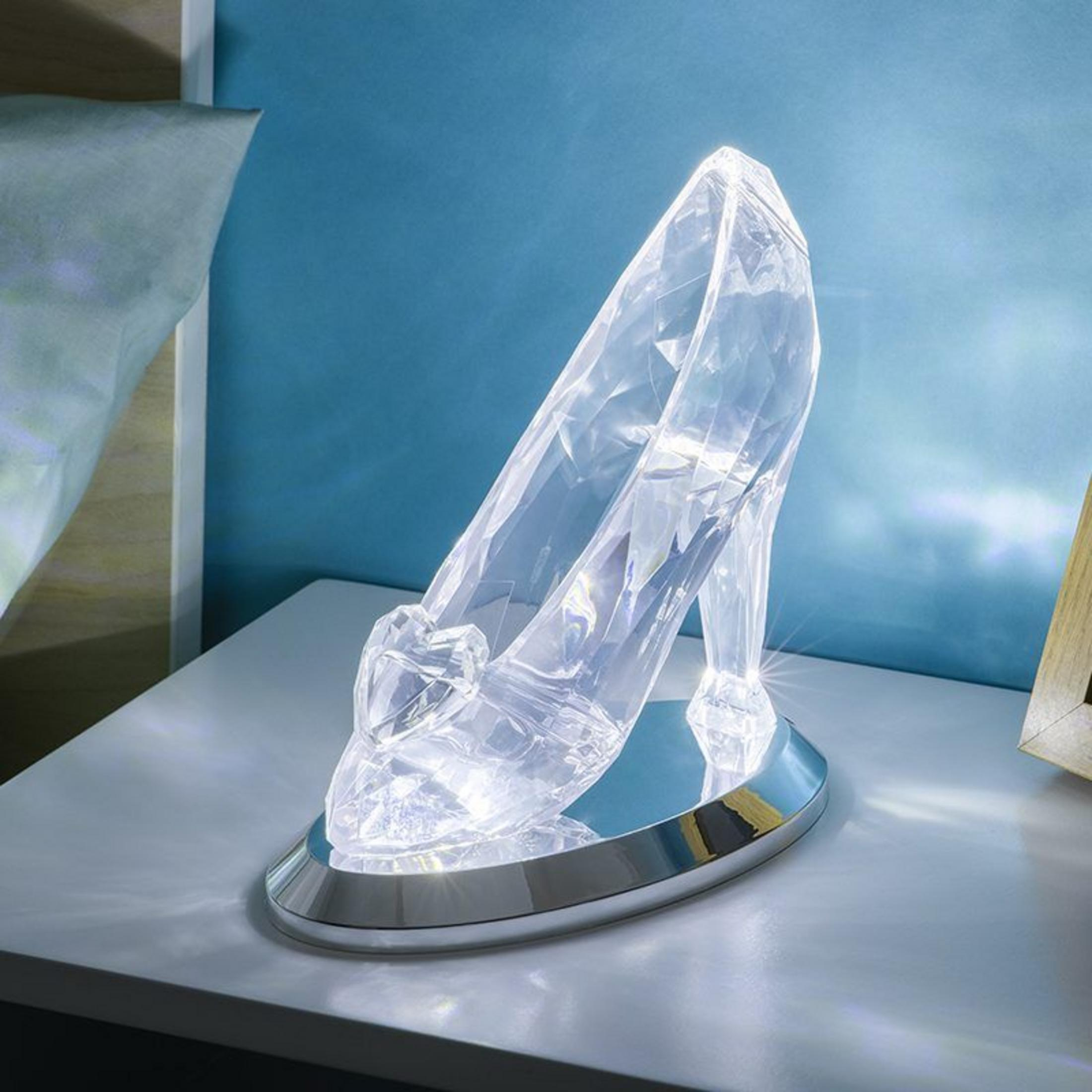 Eine durchsichtige Glas-Stiefel-Lampe mit silbernem Sockel leuchtet auf einem Nachttisch.
