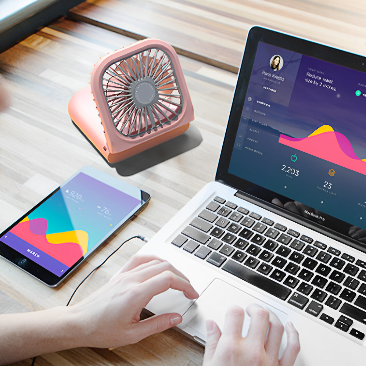 Ein pinker Ventilator, ein Tablet und ein Laptop auf einem Holztisch. Hände benutzen den Laptop.