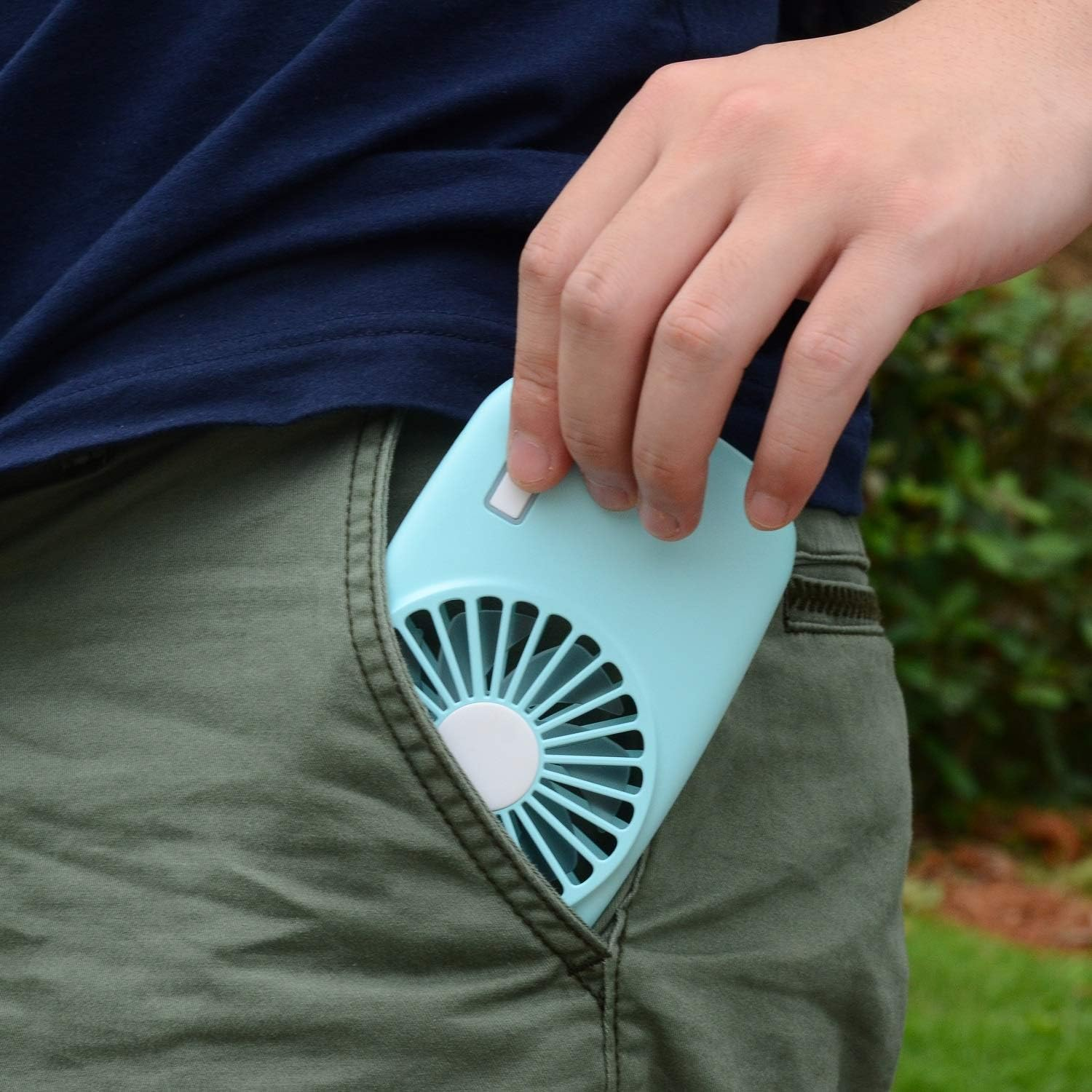 Eine Hand steckt einen hellblauen tragbaren Ventilator in die Tasche einer olivgrünen Hose.