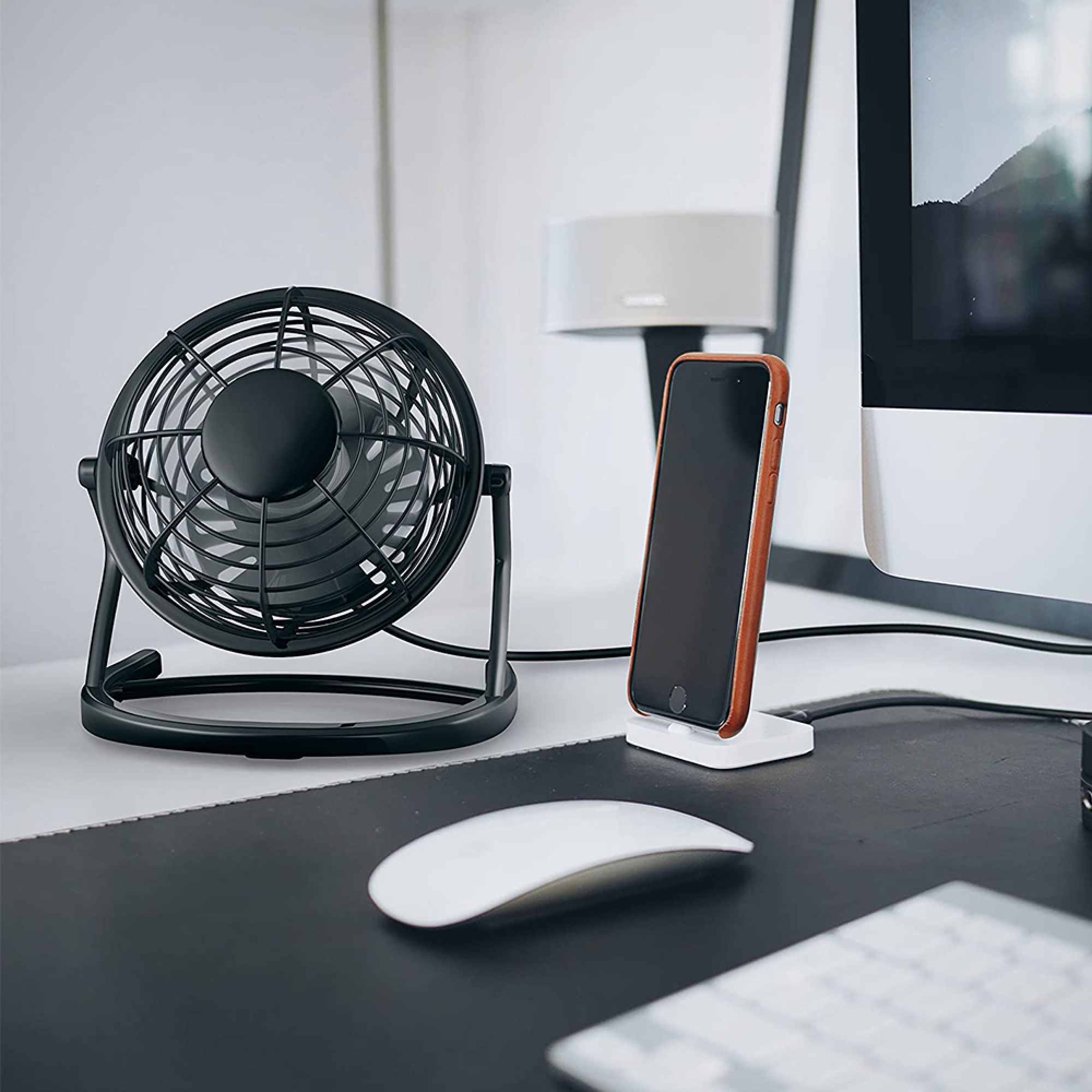 Ein schwarzer Schreibtischventilator, Handy auf Ständer, Maus und Computermonitor auf einem Schreibtisch.