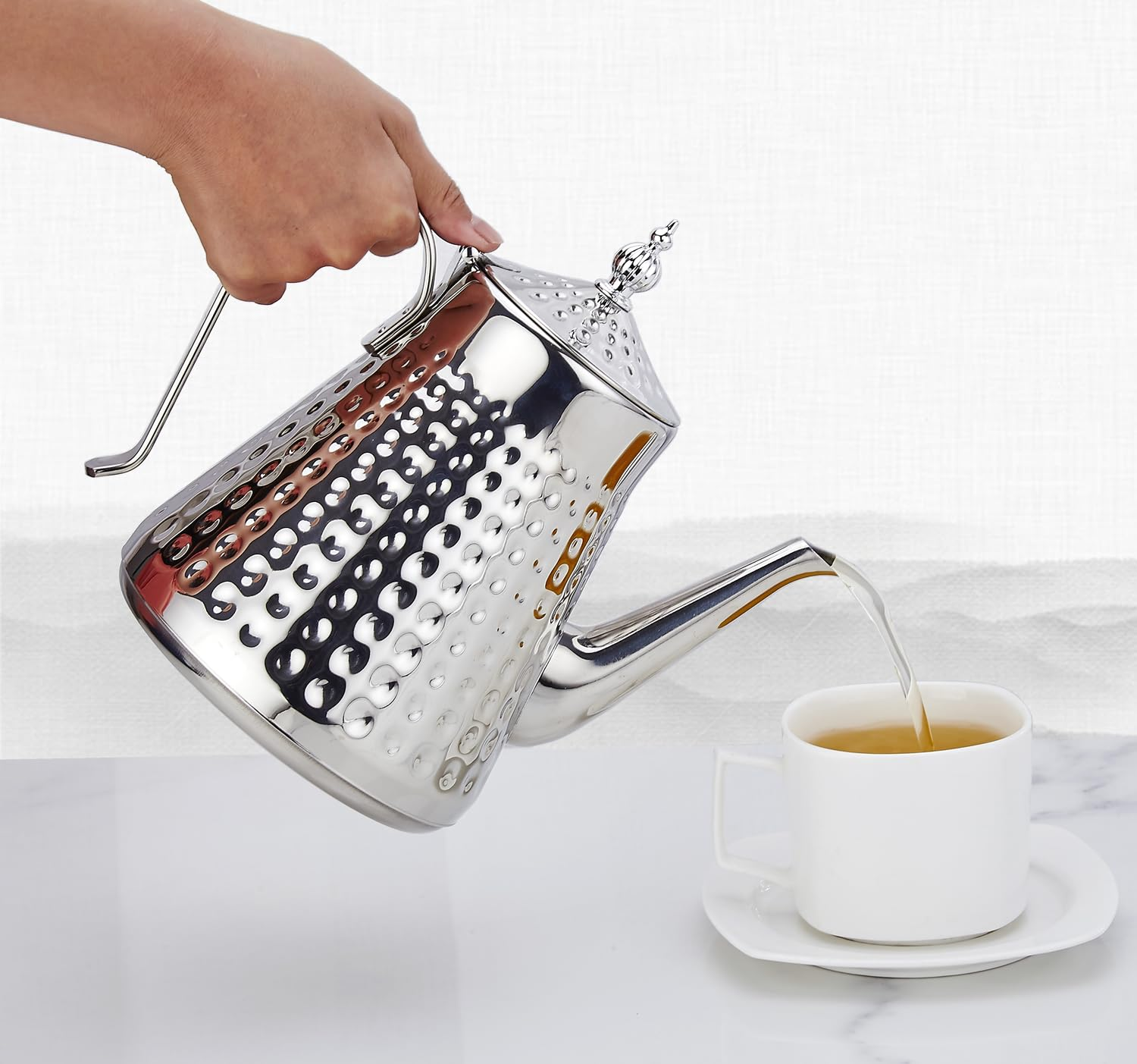 Silberne Kanne gießt Tee in weiße Tasse auf marmorierter Oberfläche.