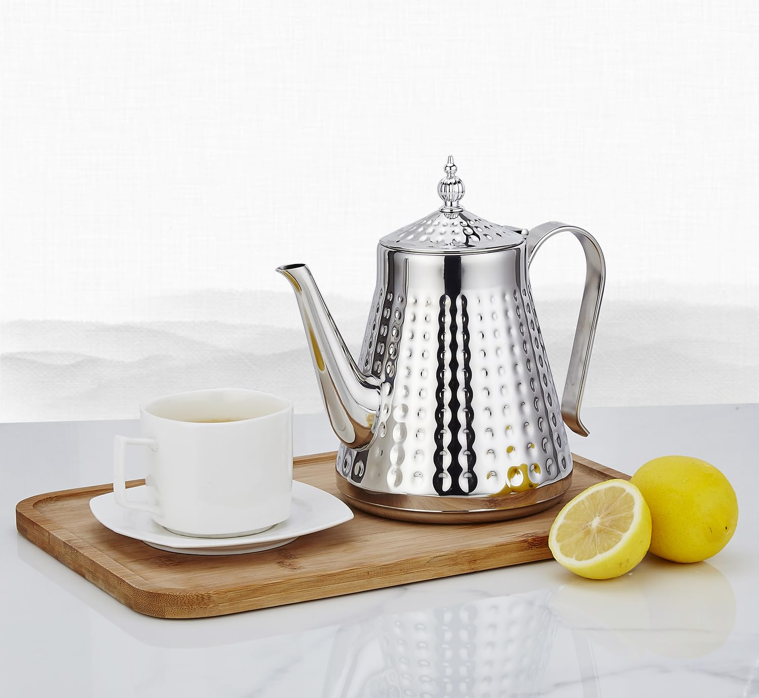 Eine silberne Teekanne mit strukturierter Oberfläche steht neben einer weißen Teetasse auf einem Bambustablett. Zwei Zitronen sind in der Nähe.