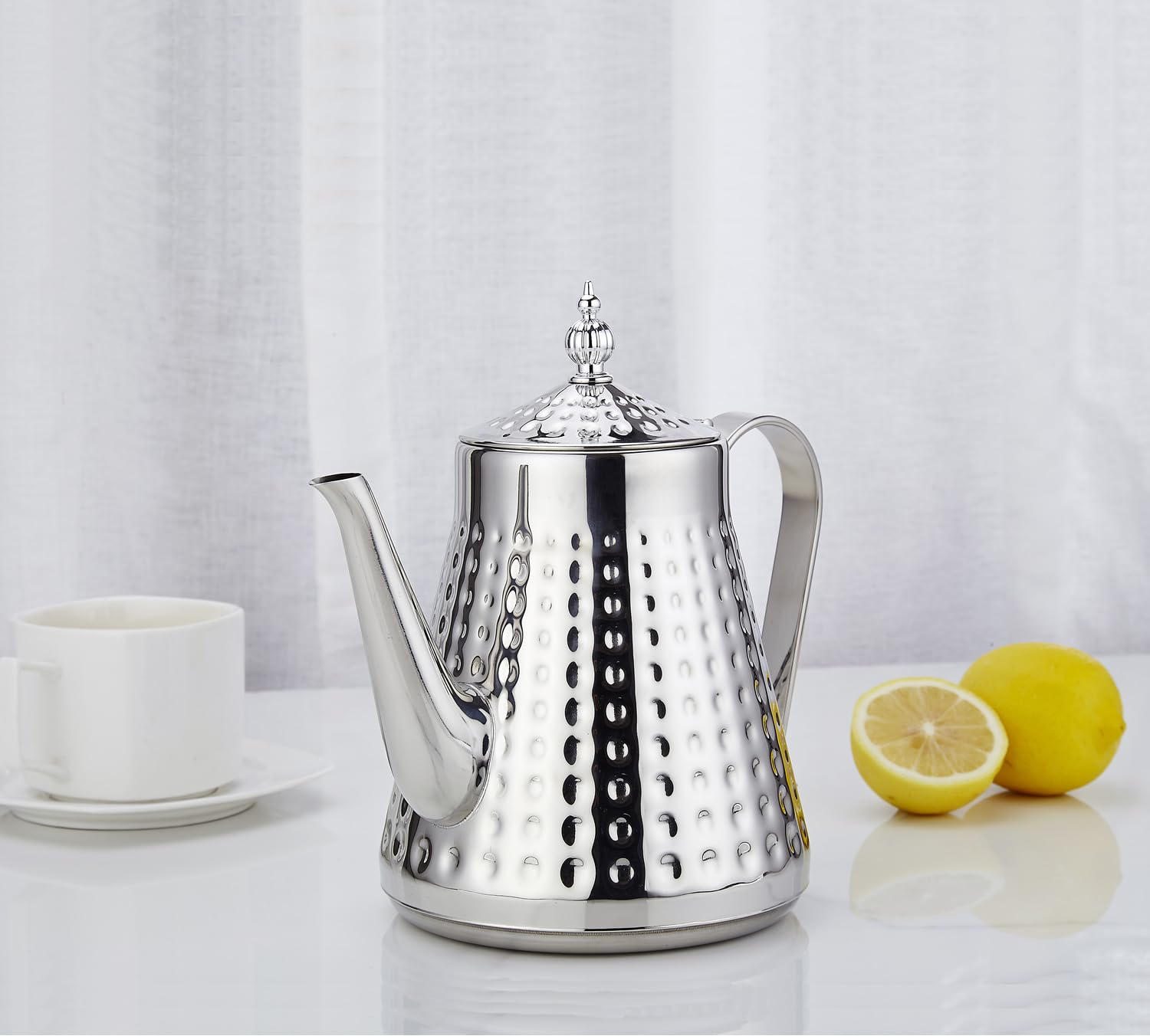 Eine silberne Teekanne mit gehämmertem Muster steht neben einer weißen Teetasse und zwei Zitronen.
