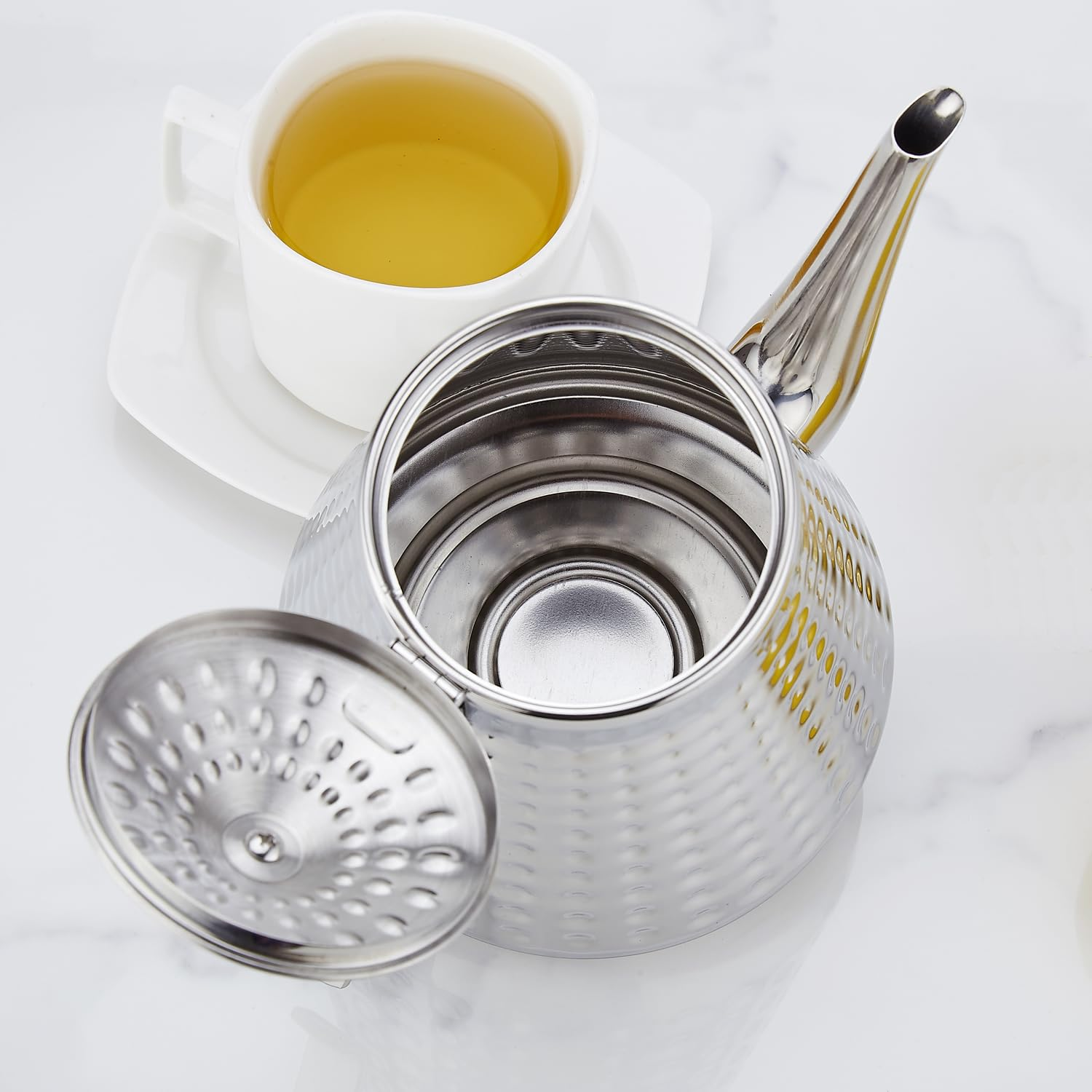 Eine silberne Teekanne mit offenem Deckel neben einer weißen Teetasse mit gelbem Tee.