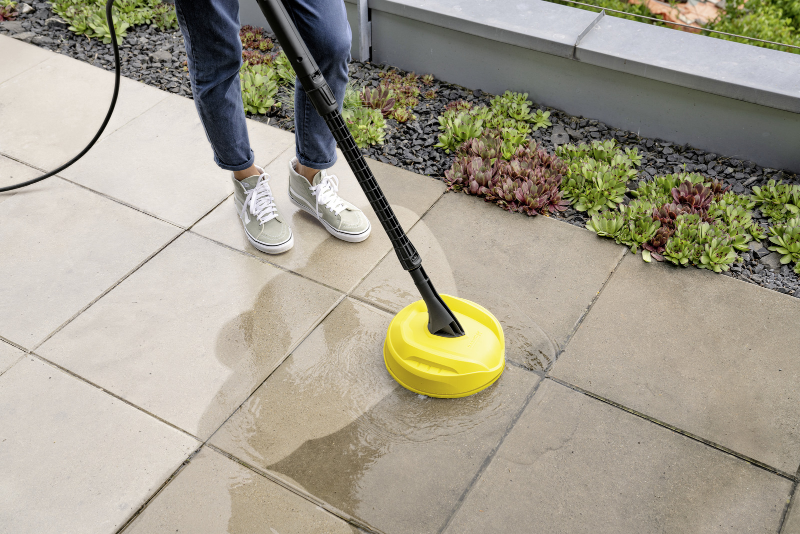 Person cleaning patio tiles with a yellow pressure washer attachment.
