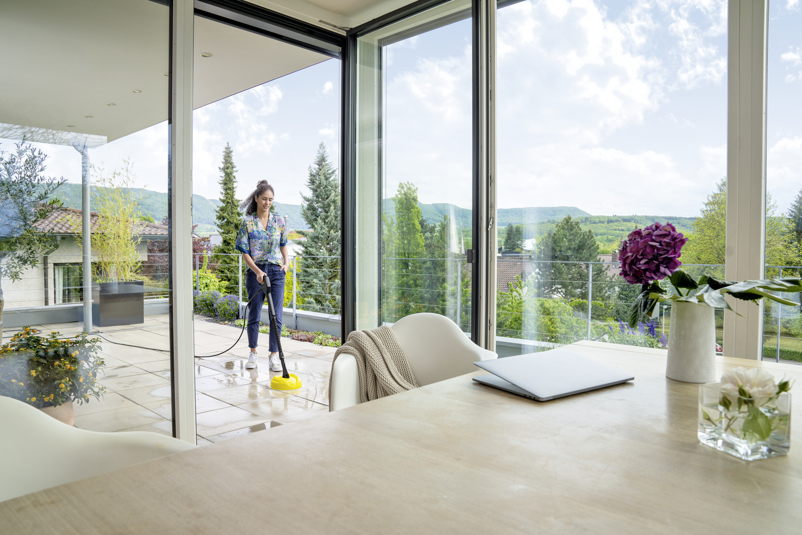 Eine Frau reinigt einen Terrassenboden mit einem gelben Hochdruckreiniger. Draußen ist eine Landschaft zu sehen.