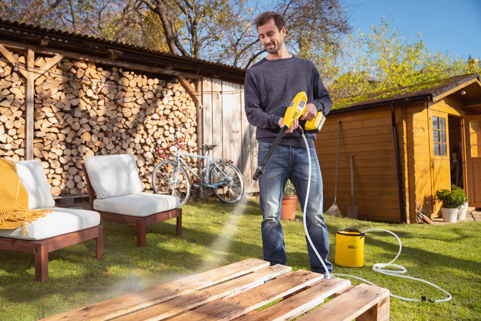 Man cleans wooden pallets with a pressure washer in the garden. Woodpile and shed in background.