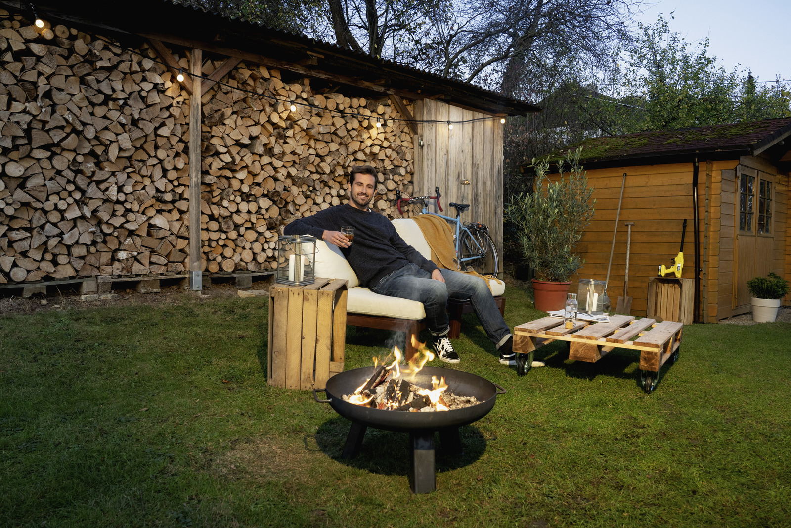 Man relaxing by a fire pit in a backyard with woodpile and shed.