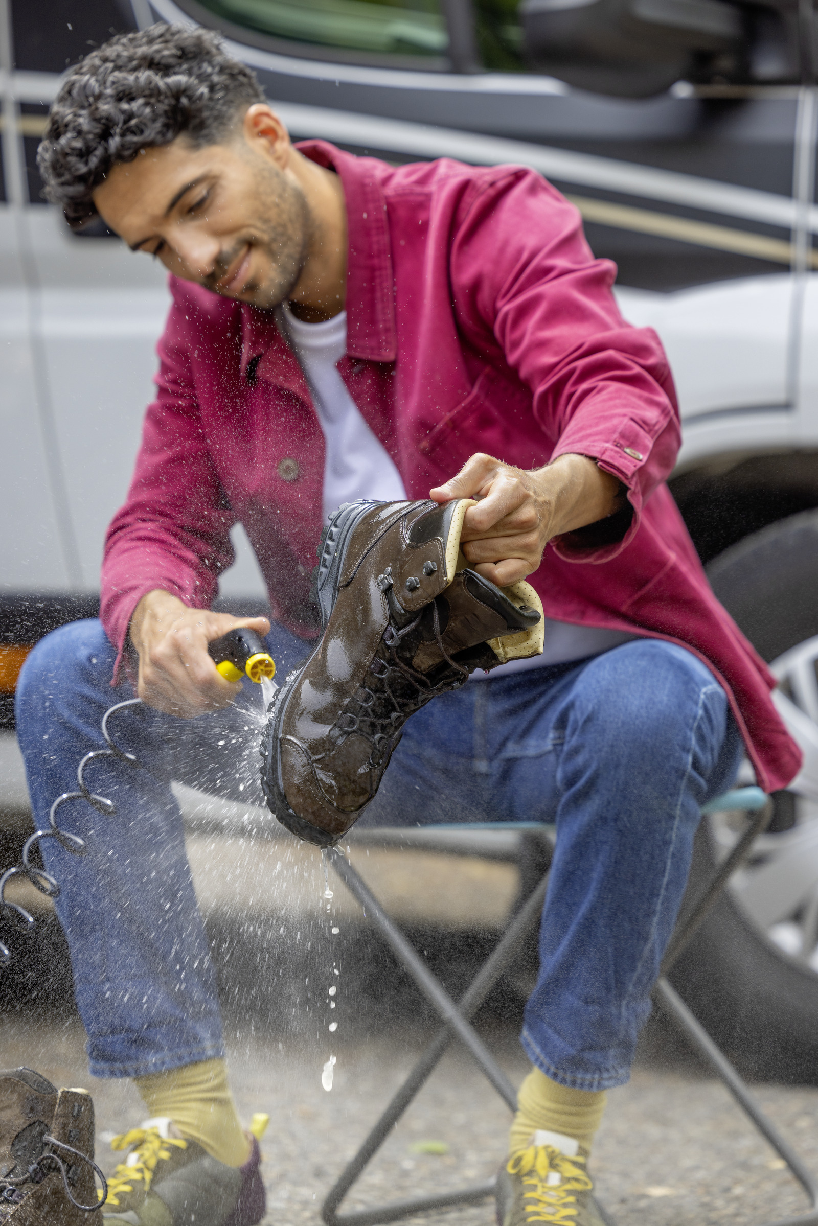 Mann reinigt braunen Stiefel mit Wassersprühpistole neben einem weißen Fahrzeug.