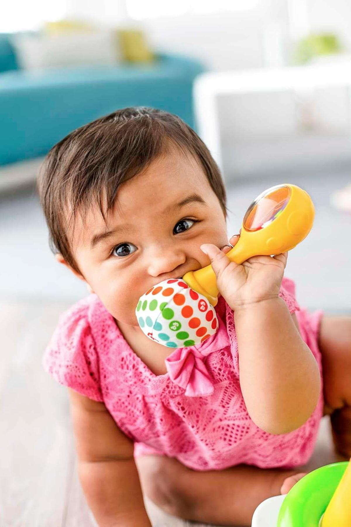 Ein Baby in einem pinken Hemd kaut auf einem bunten Tupfenspielzeug vor einer unscharfen blauen Couch.