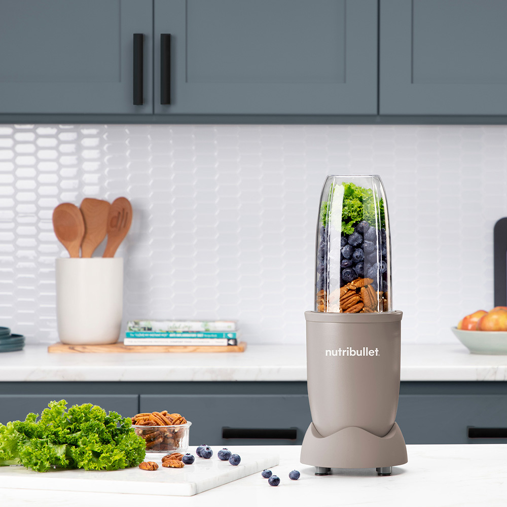 A Nutribullet blender with blueberries, nuts, and lettuce. Ingredients on marble board.