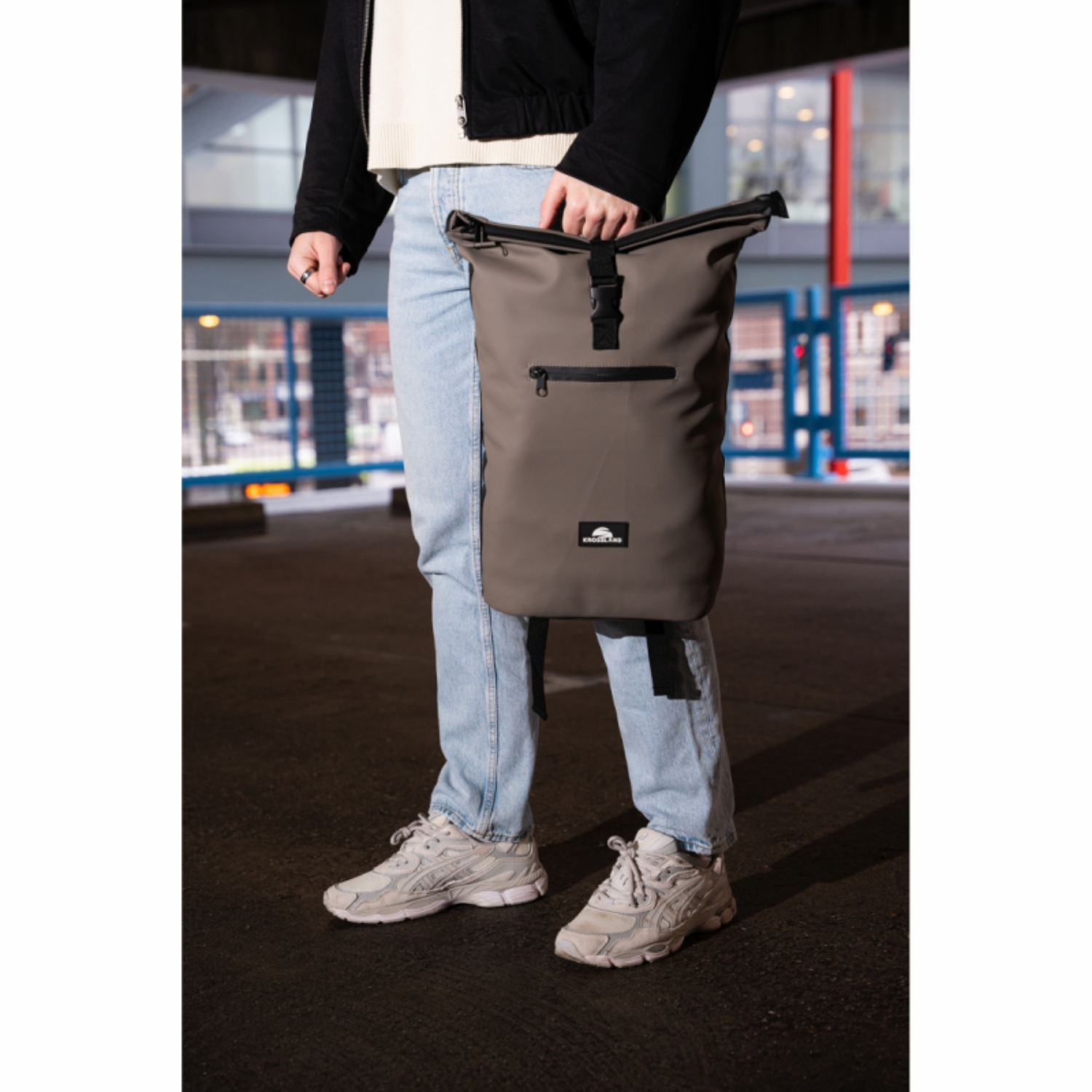 Een persoon in jeans en sneakers draagt een grijze roltop-tas.