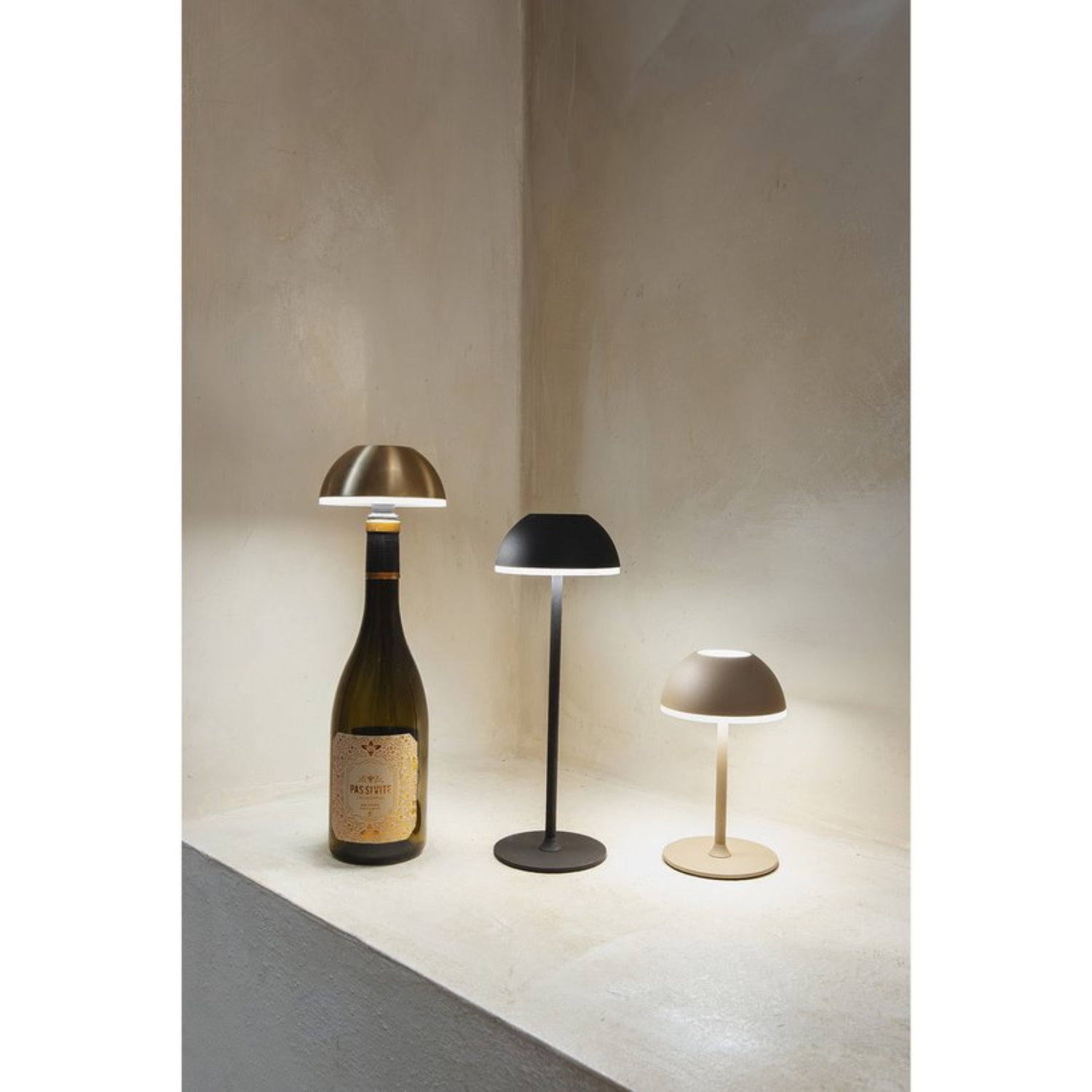 Three table lamps on a shelf: a gold one above a wine bottle, a black one, and a beige one.