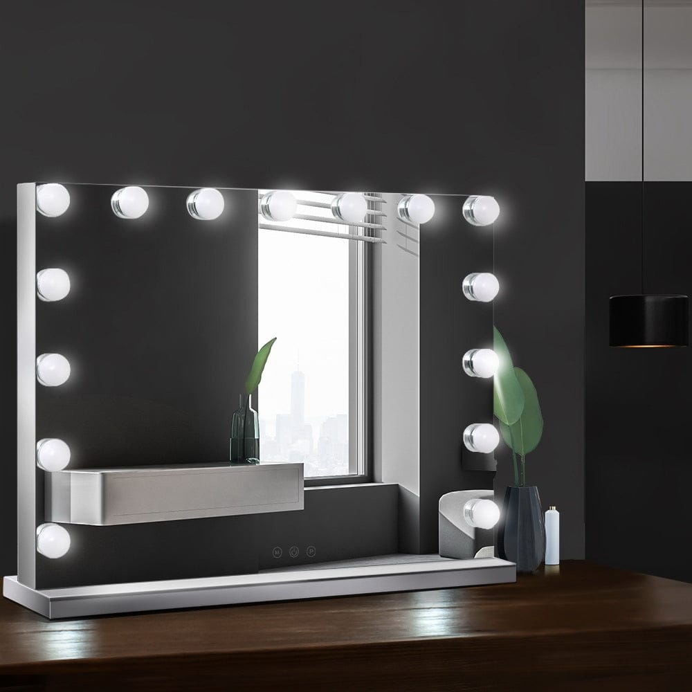 Uno specchio per il trucco illuminato con lampadine e un ripiano si trova su un tavolo di legno.