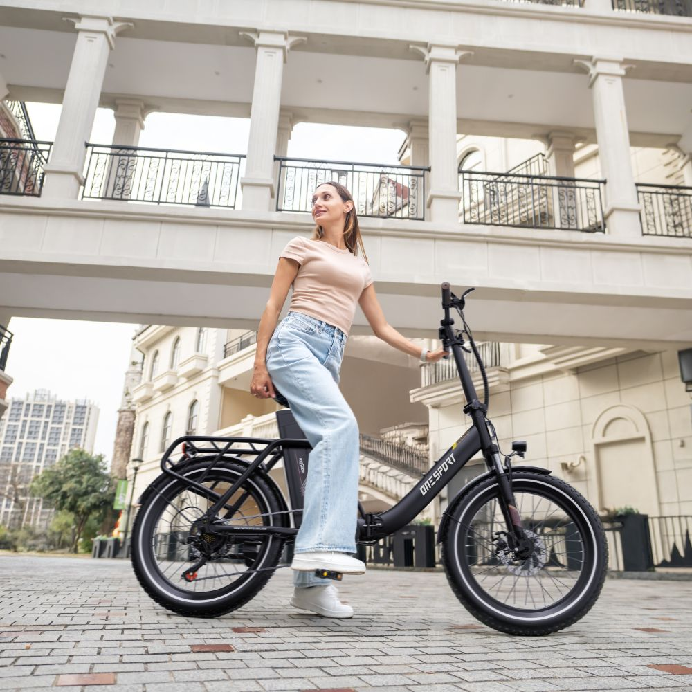 Una donna con maglietta color cuoio e jeans chiari guida una bici elettrica nera su strada pavimentata.