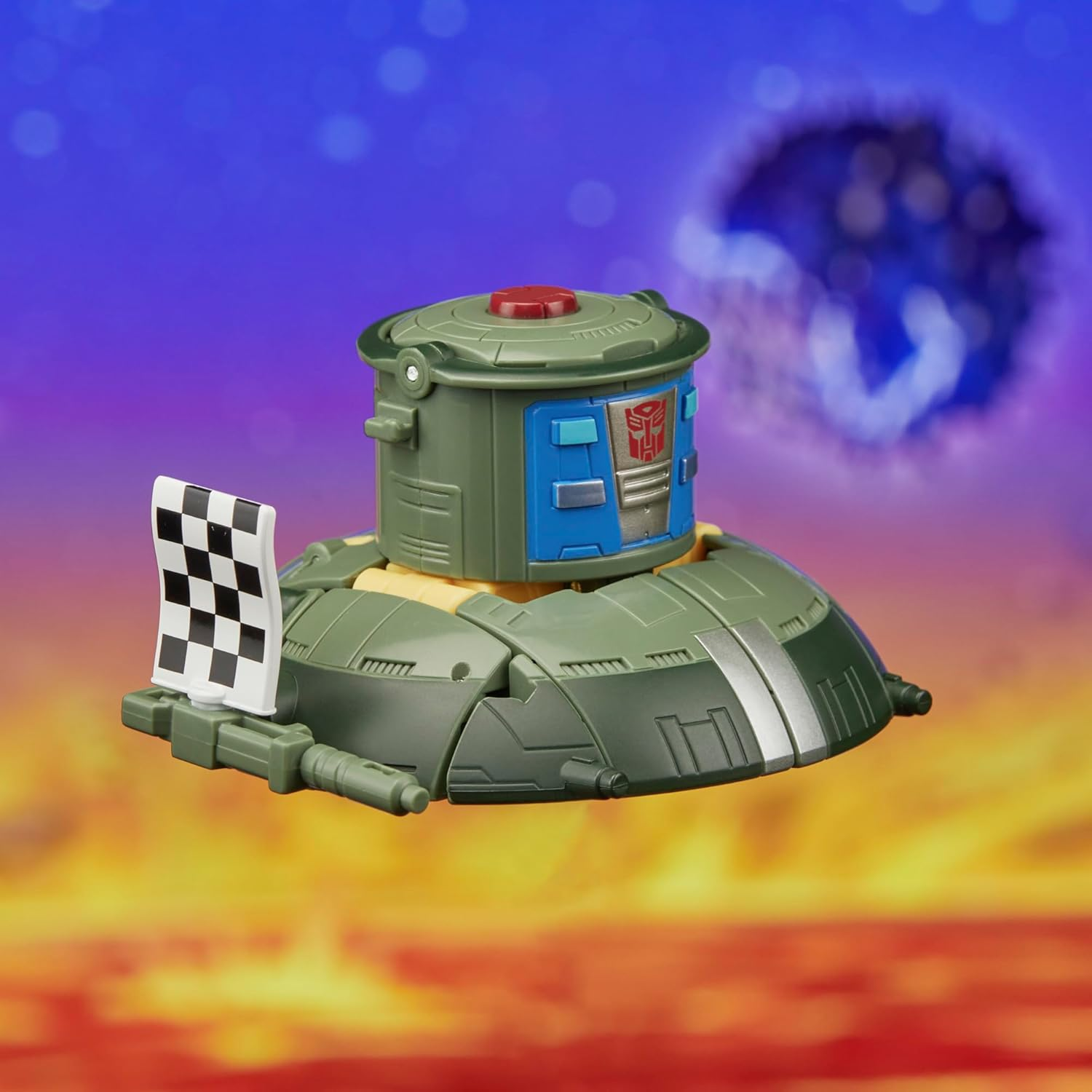 Zielono-niebieska zabawka Transformer z flagą w kratkę na tle fioletowo-pomarańczowego nieba.