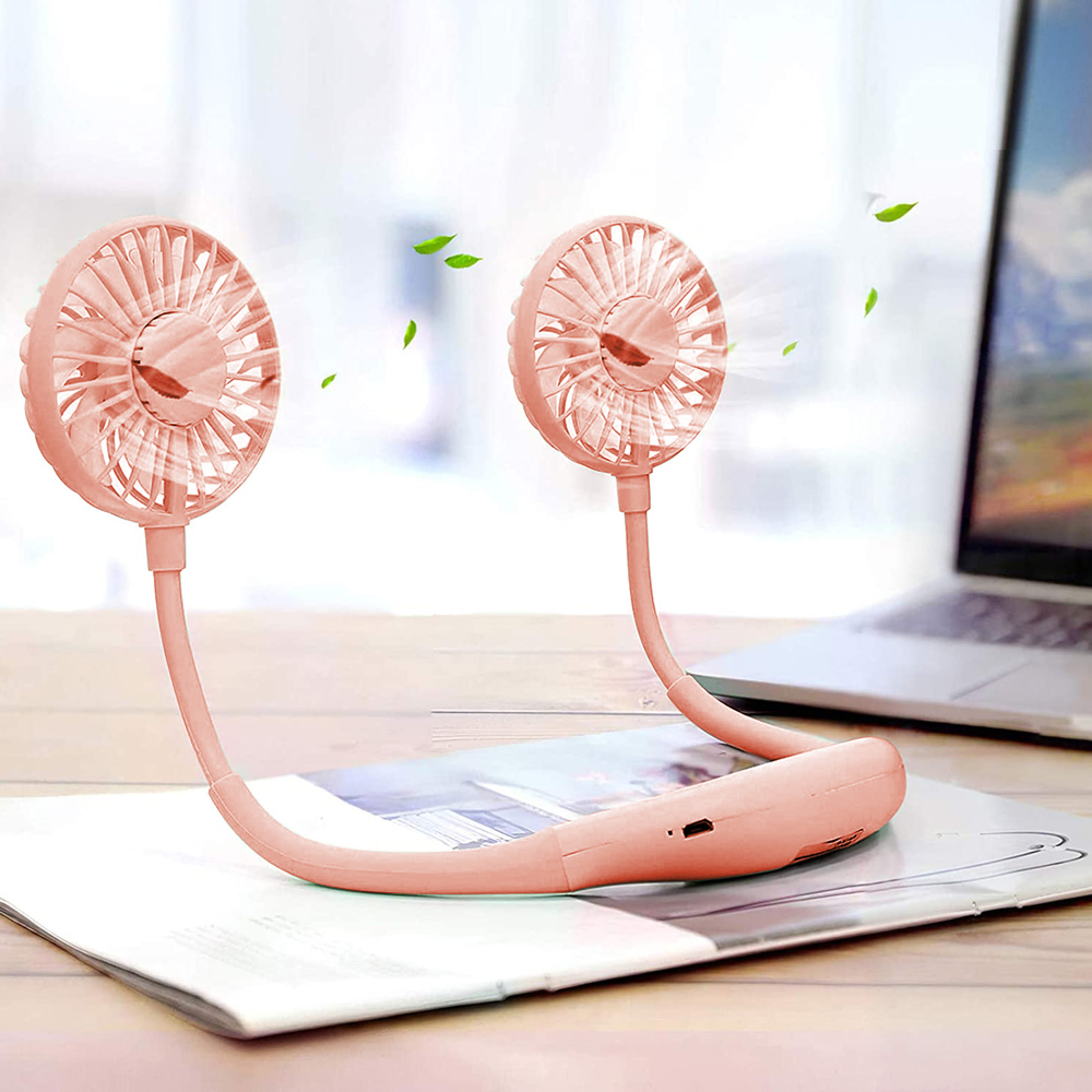 Ein rosafarbener Nackenventilator mit zwei Lüfterköpfen liegt auf einem Magazin neben einem Laptop.