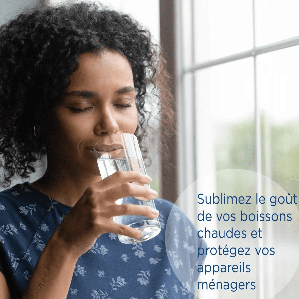 Eine Frau mit lockigem Haar trinkt aus einem Glas Wasser mit Blick auf ein Fenster.