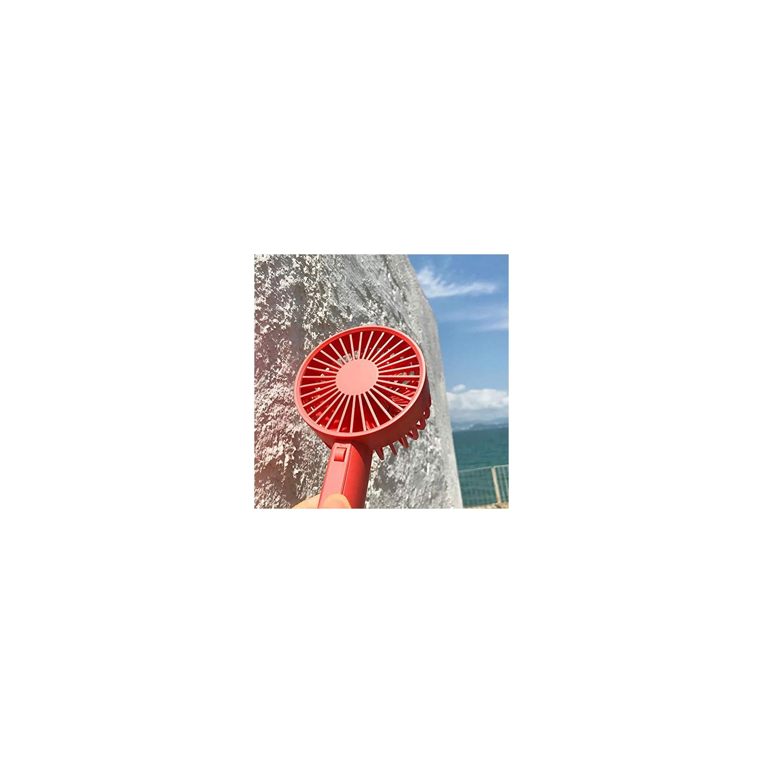 Ein roter tragbarer Ventilator wird gegen eine strukturierte Wand gehalten. Der Hintergrund zeigt das Meer und den Himmel.