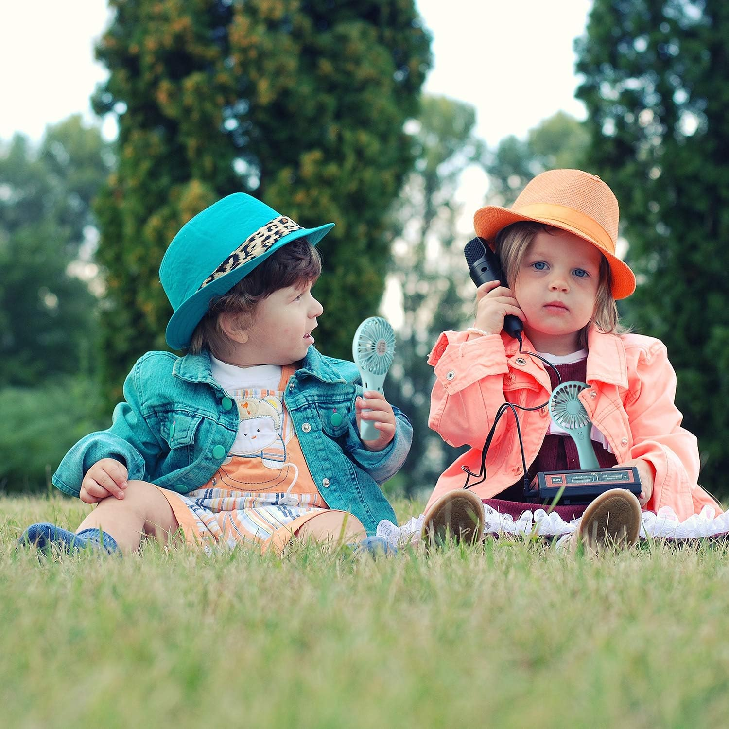 Zwei Kinder sitzen im Gras. Eines hält ein Telefon, das andere einen Ventilator.