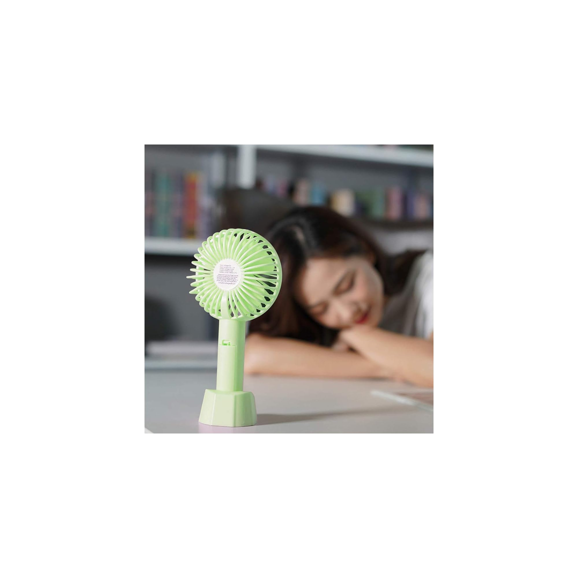 Ein kleiner, heller Ventilator steht auf einem Tisch, eine Frau schläft im Hintergrund.