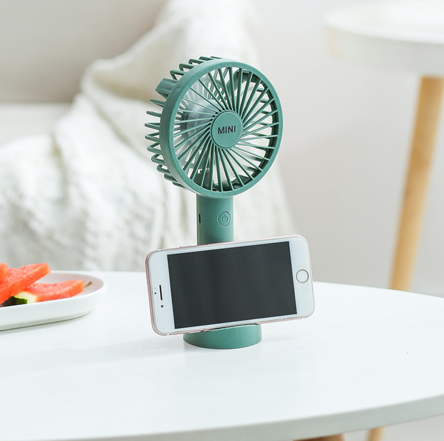 Ein grüner Mini-Ventilator hält ein Smartphone. Wassermelonenstücke sind im Hintergrund.