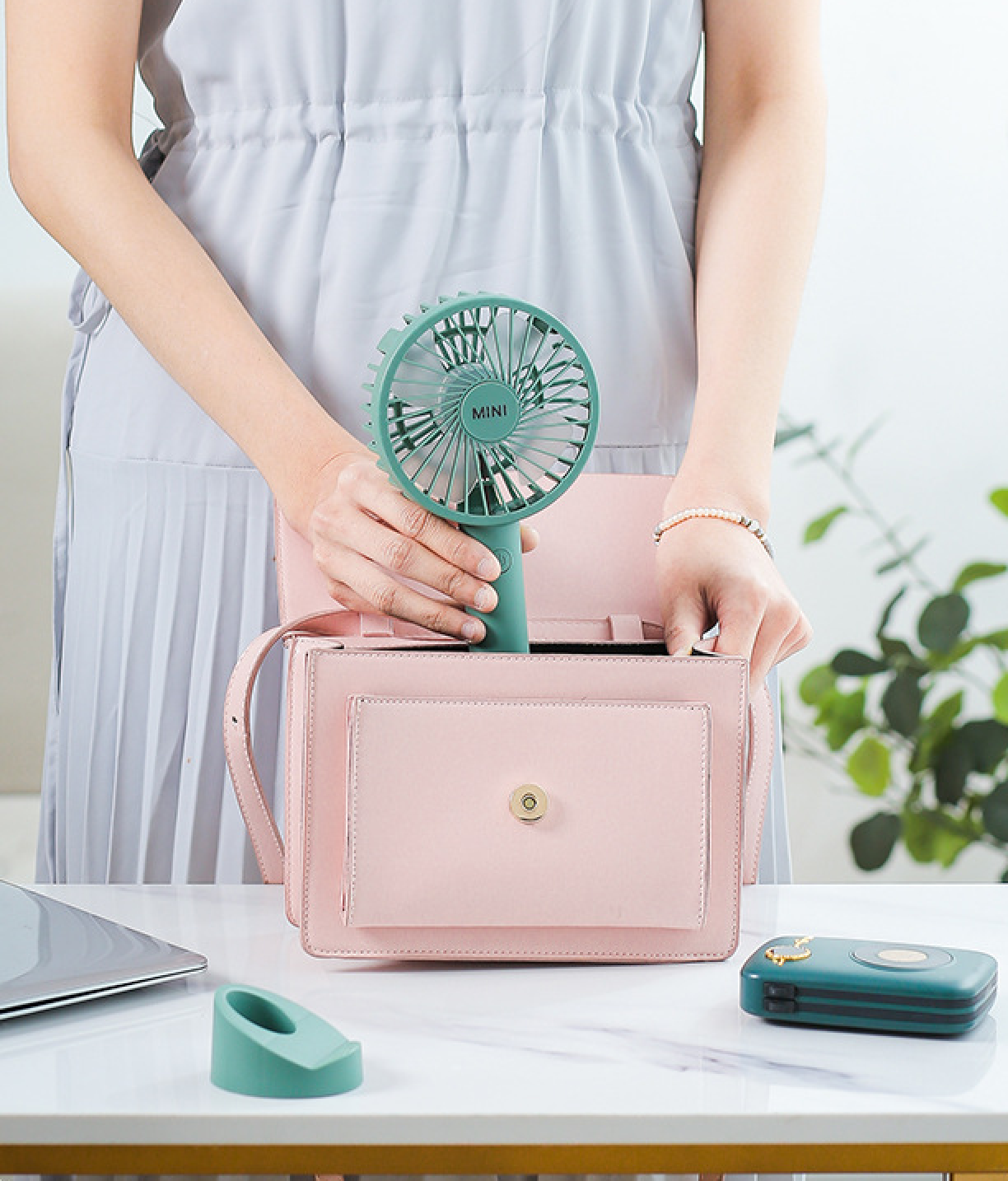 Eine Person steckt einen grünen Mini-Ventilator in eine rosa Handtasche. Ein Laptop und ein Ventilatorständer sind in der Nähe.
